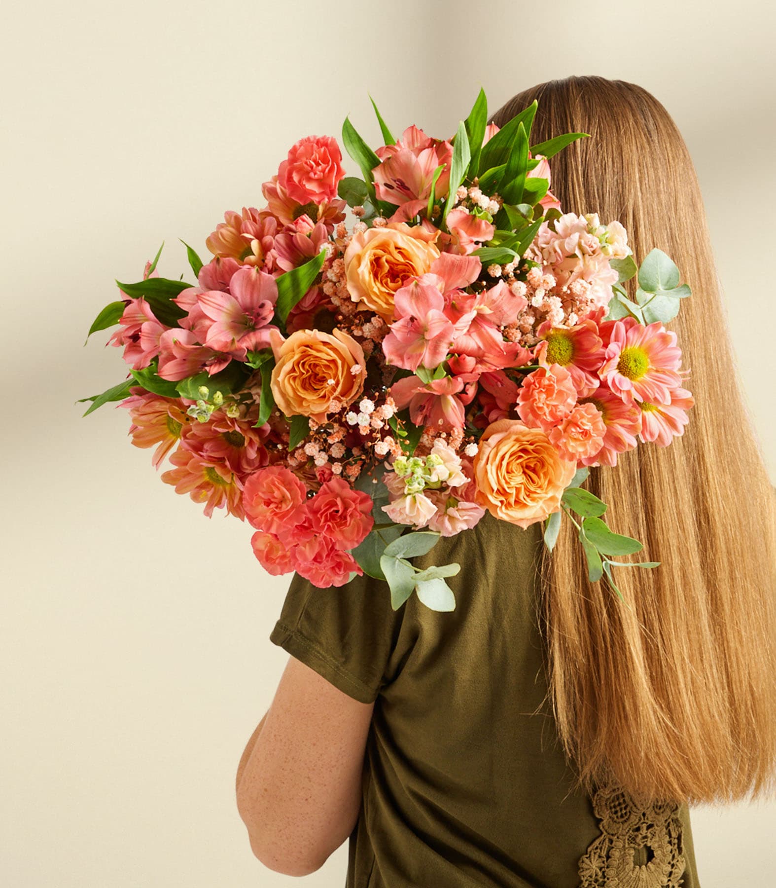 Ein üppiger Strauß bunter Blumen, darunter Rosen, Nelken und andere Blüten, wird vor einer Person mit langen, blonden Haaren vor einem schlichten, hellen Hintergrund gehalten.