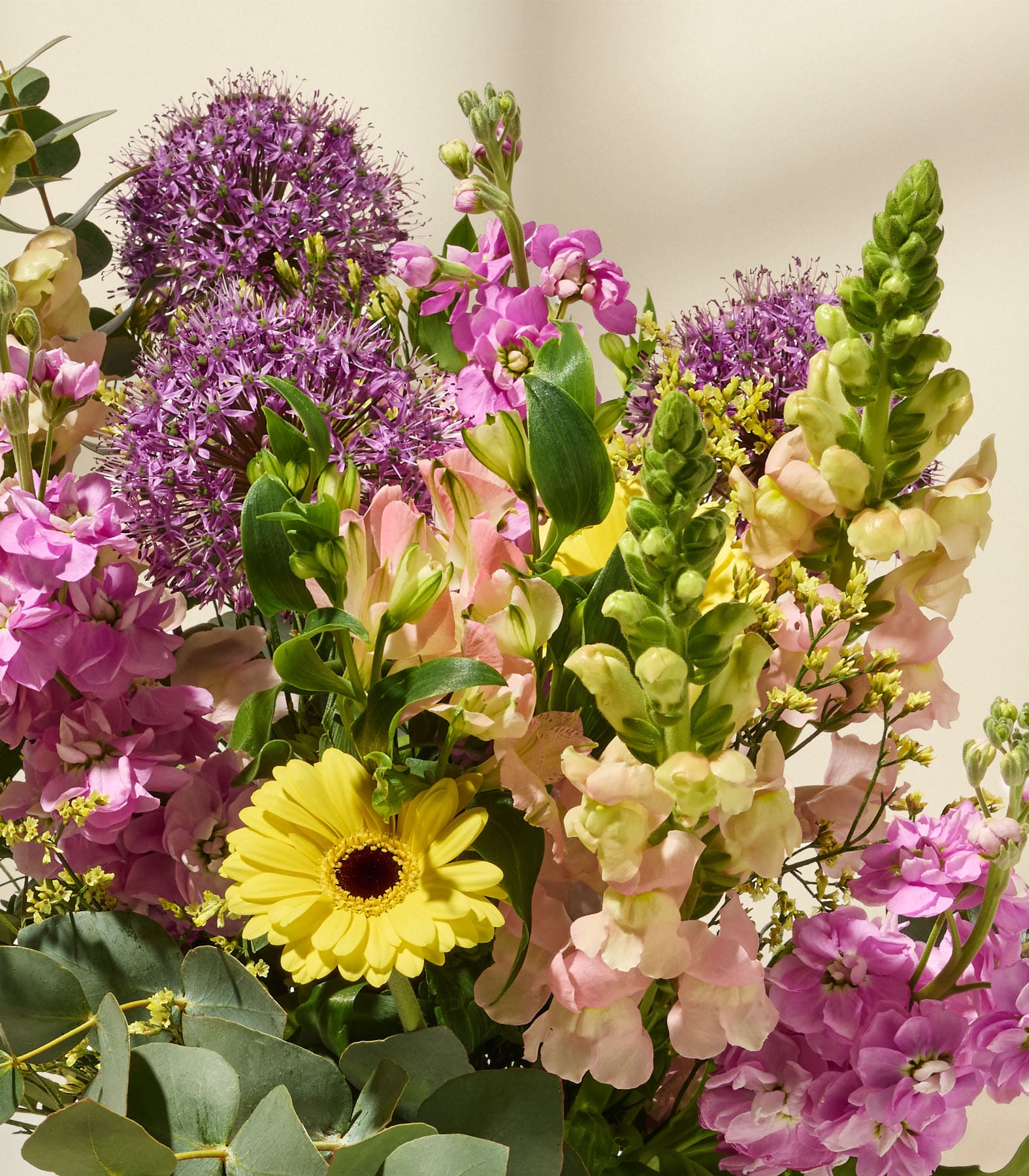 Ein lebhafter Strauß aus verschiedenen farbenfrohen Blumen, darunter lila Zierlauch, rosa Löwenmäulchen und gelbe Gerbera, vor einem neutralen Hintergrund.