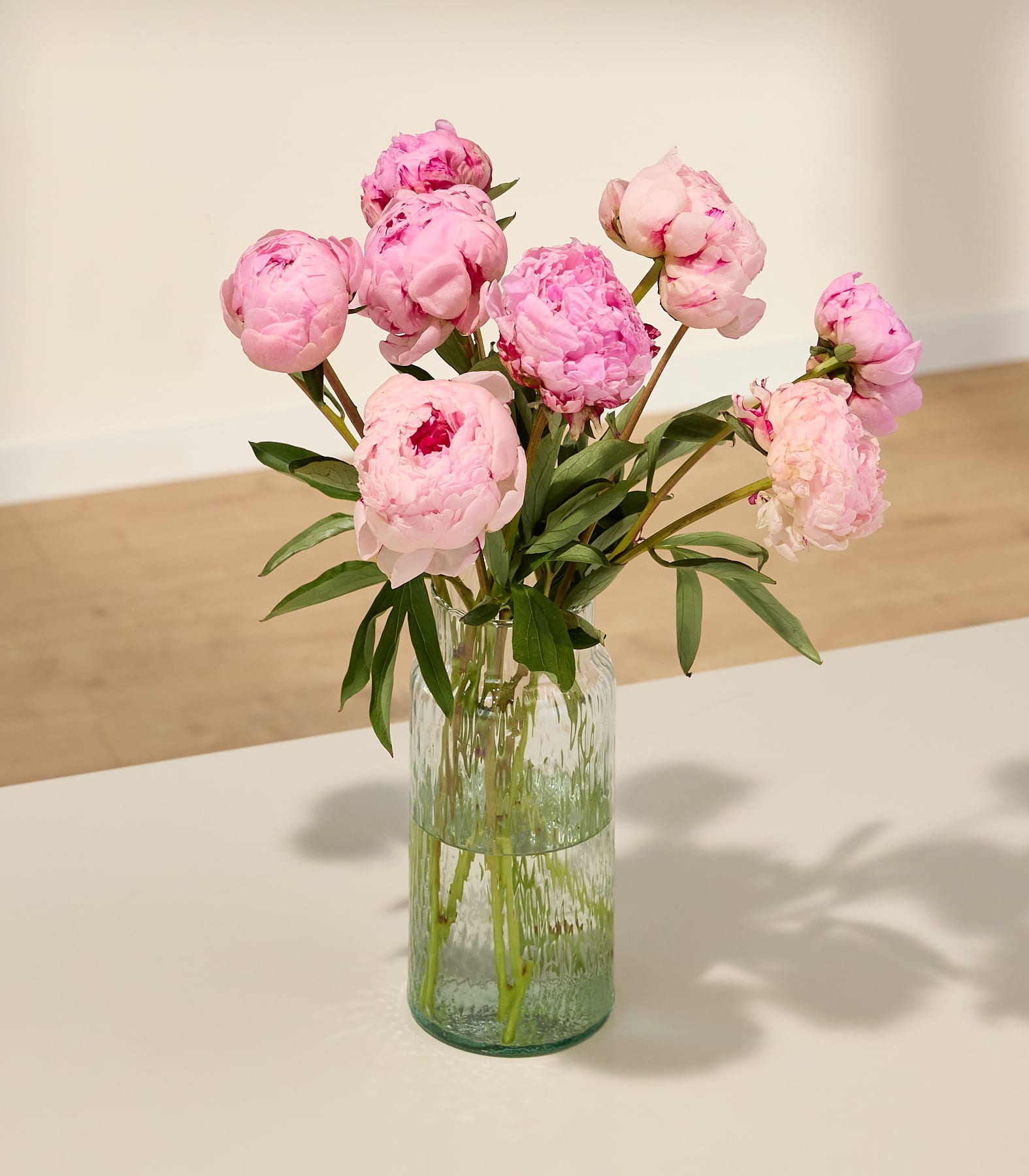 Ein Strauß leuchtend pinkfarbener Pfingstrosen in einer Glasvase steht auf einer Holzoberfläche; der schlichte Hintergrund sorgt für eine einfache und elegante Atmosphäre.