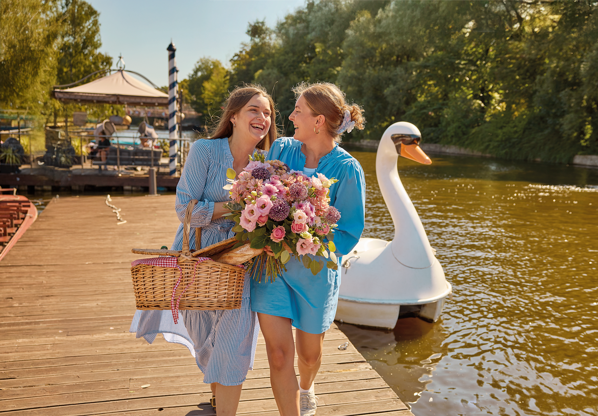 Zwei Frauen in legerer Sommerkleidung spazieren gemeinsam auf einem Holzsteg. Sie tragen einen Blumenstrauß und einen Picknickkorb. Hinter ihnen schwimmt ein Schwan im Wasser, und im Hintergrund ist ein Pavillon zu sehen.