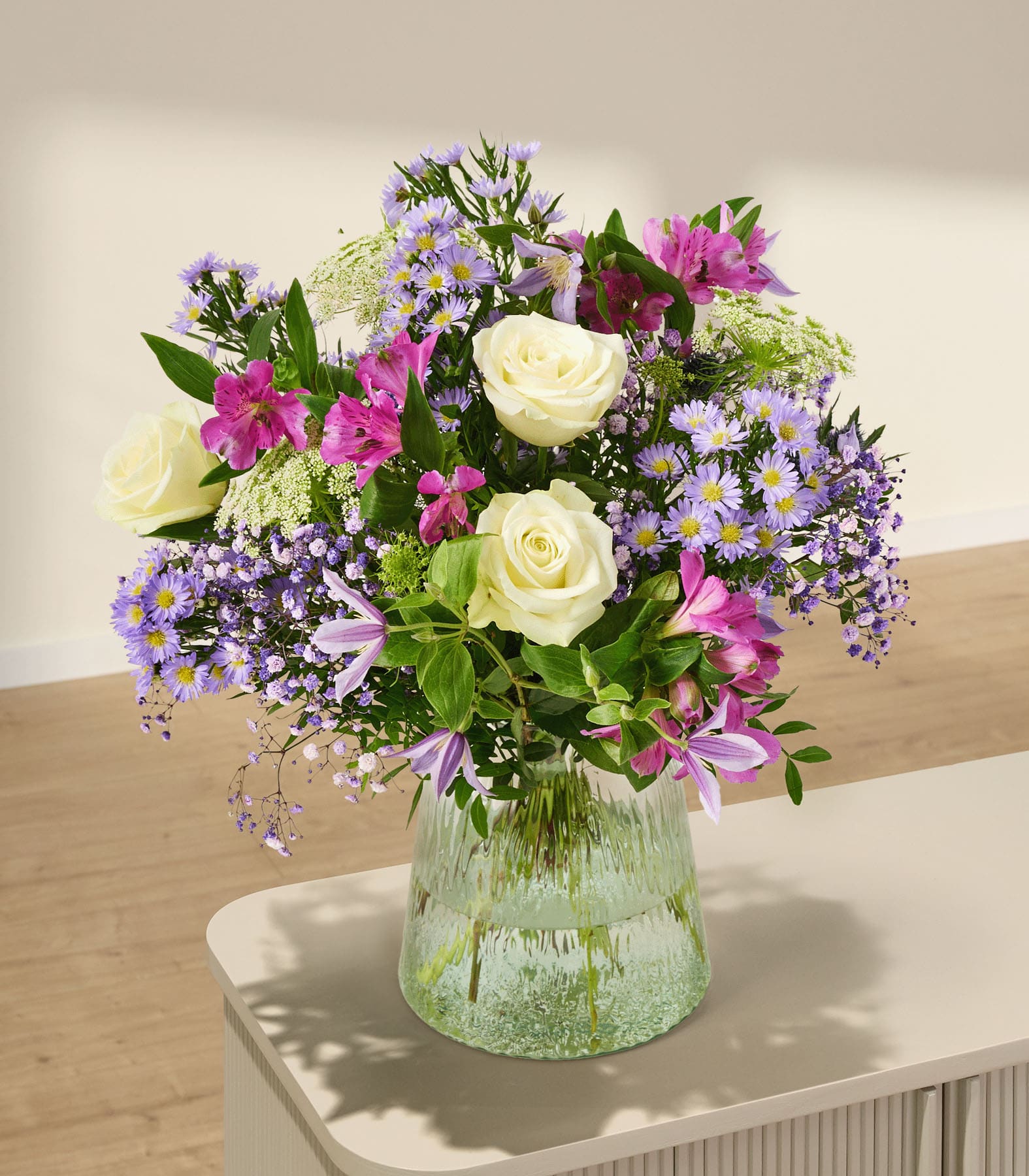Ein leuchtender Strauß verschiedener bunter Blumen, darunter Rosen, Gänseblümchen und Flieder, den eine Person vor einem schlichten weißen Hintergrund in den Händen hält.