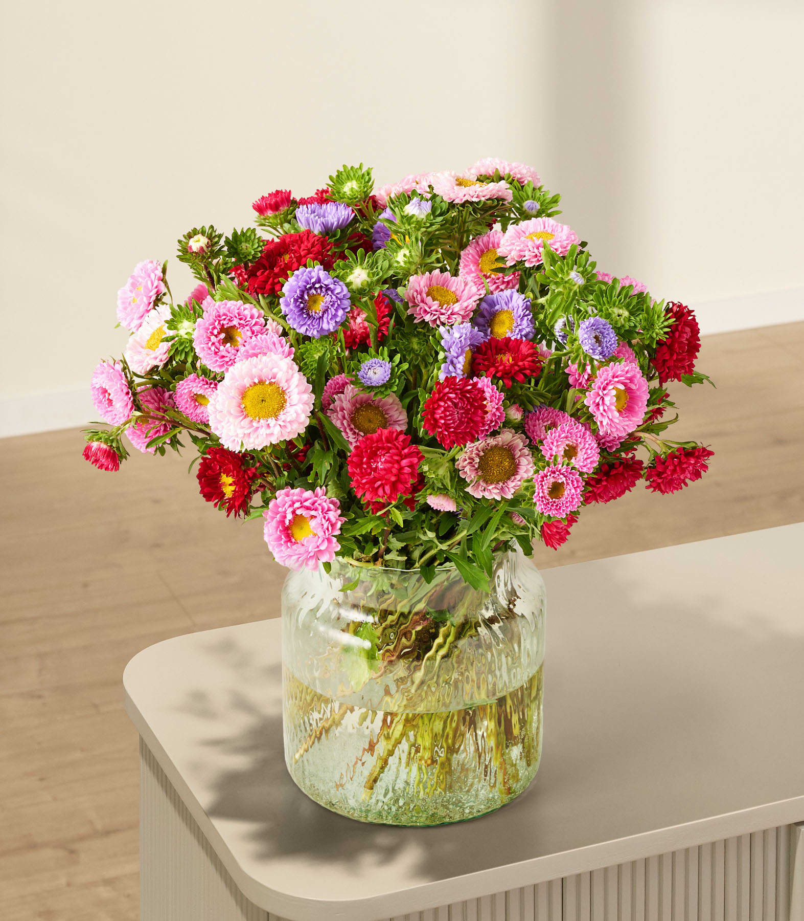 Ein leuchtender Strauß bunter Blumen, darunter rosa und rote Dahlien, weiße Gänseblümchen und grünes Laub, wird vor einem schlichten weißen Hintergrund in den Händen einer Person gehalten.