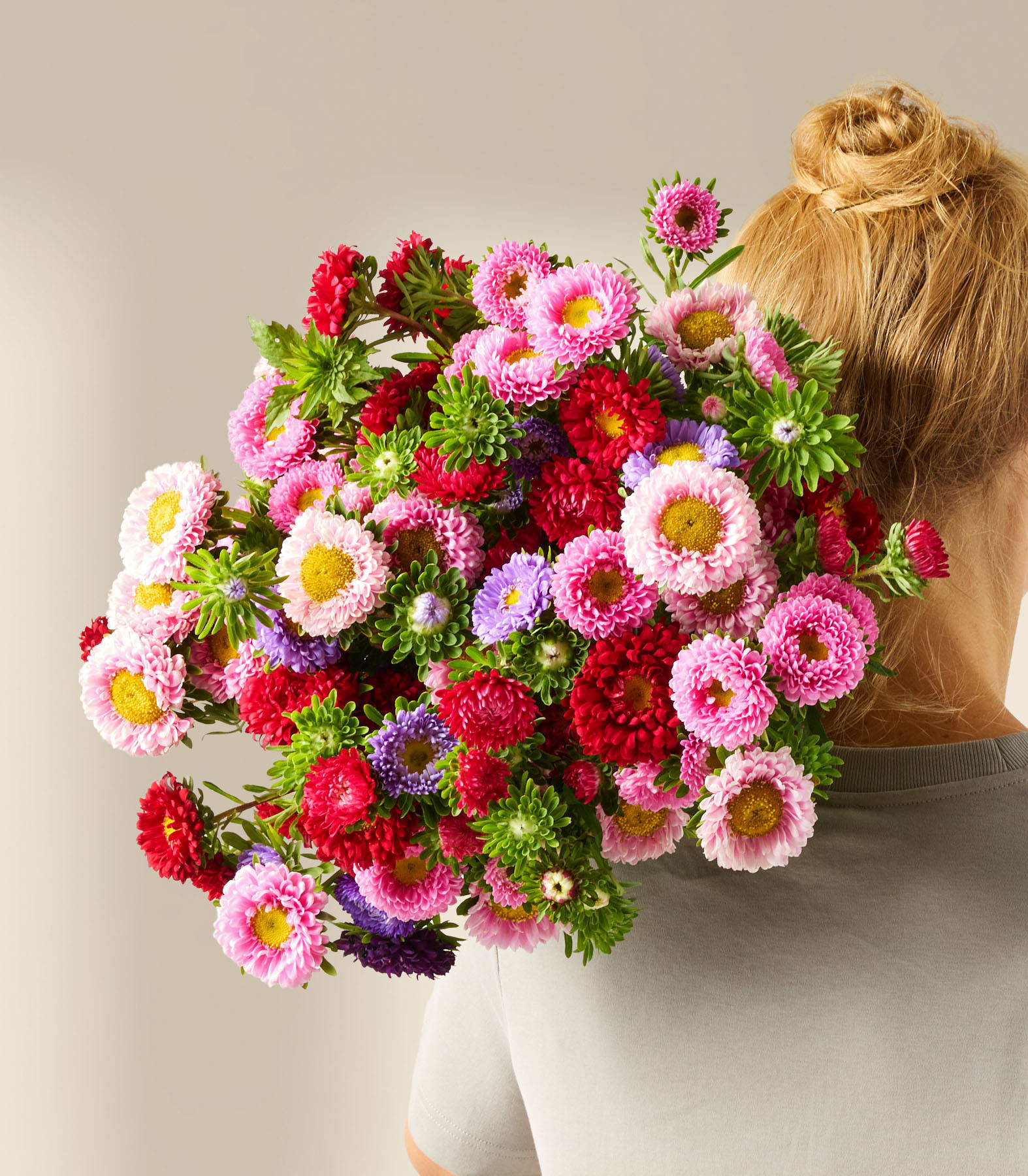 Ein leuchtender Strauß bunter, gänseblümchenartiger Blumen in Rosa-, Rot- und Lilatönen wird in den Händen einer Person gehalten, die ein graues Hemd vor einem schlichten Hintergrund trägt.