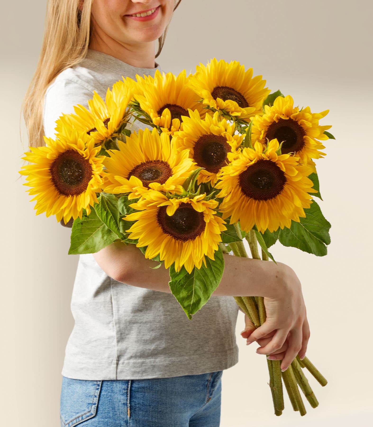 Eine lächelnde Frau hält einen Strauß leuchtend gelber Sonnenblumen vor einem schlichten weißen Hintergrund.