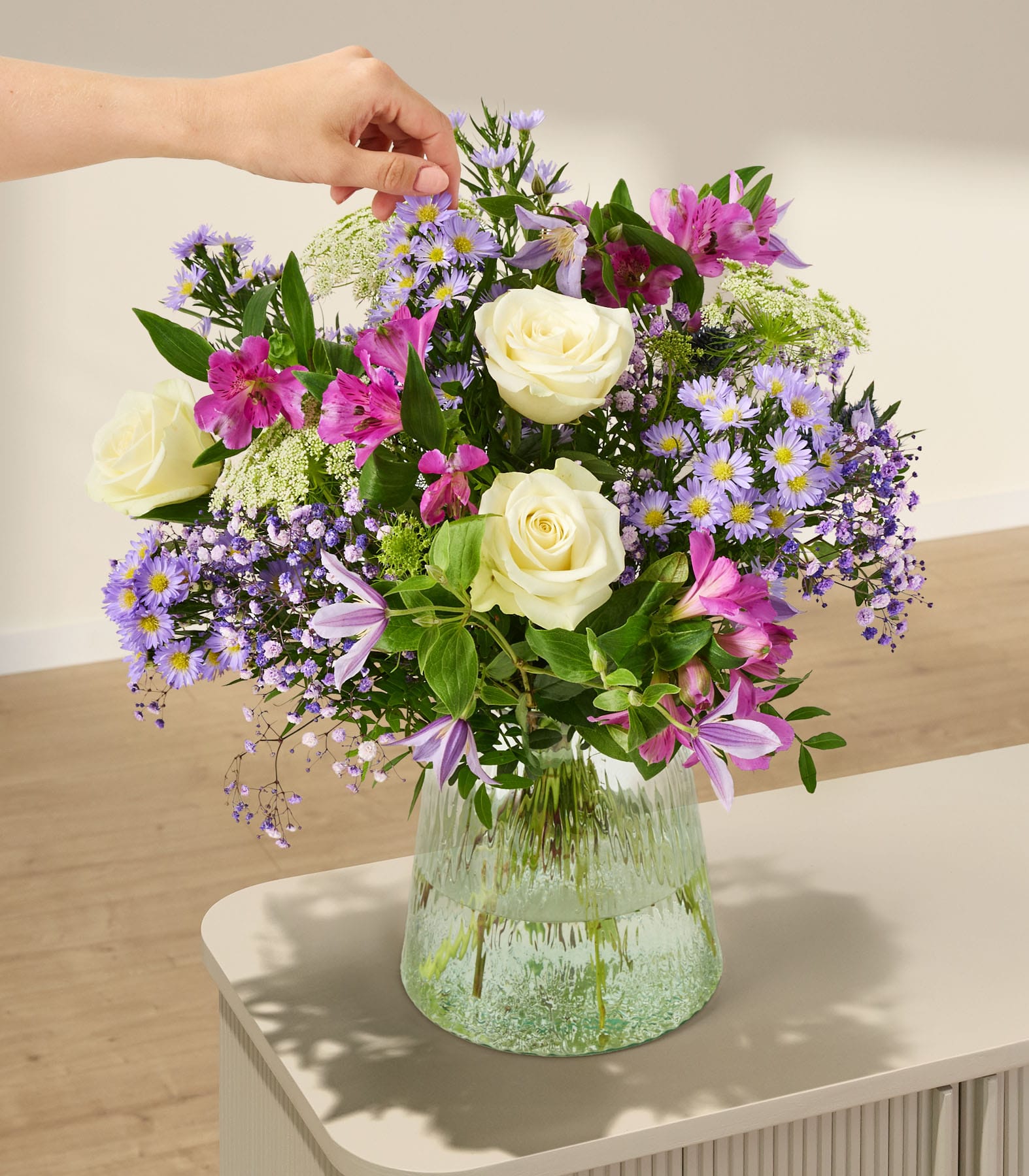 Ein farbenfroher Strauß verschiedener Blumen, darunter weiße Rosen, violette Gänseblümchen und rosa Lilien, wird in einer Glasvase vor einem schlichten weißen Hintergrund präsentiert, und eine Hand greift hinein, um das Arrangement zu richten.