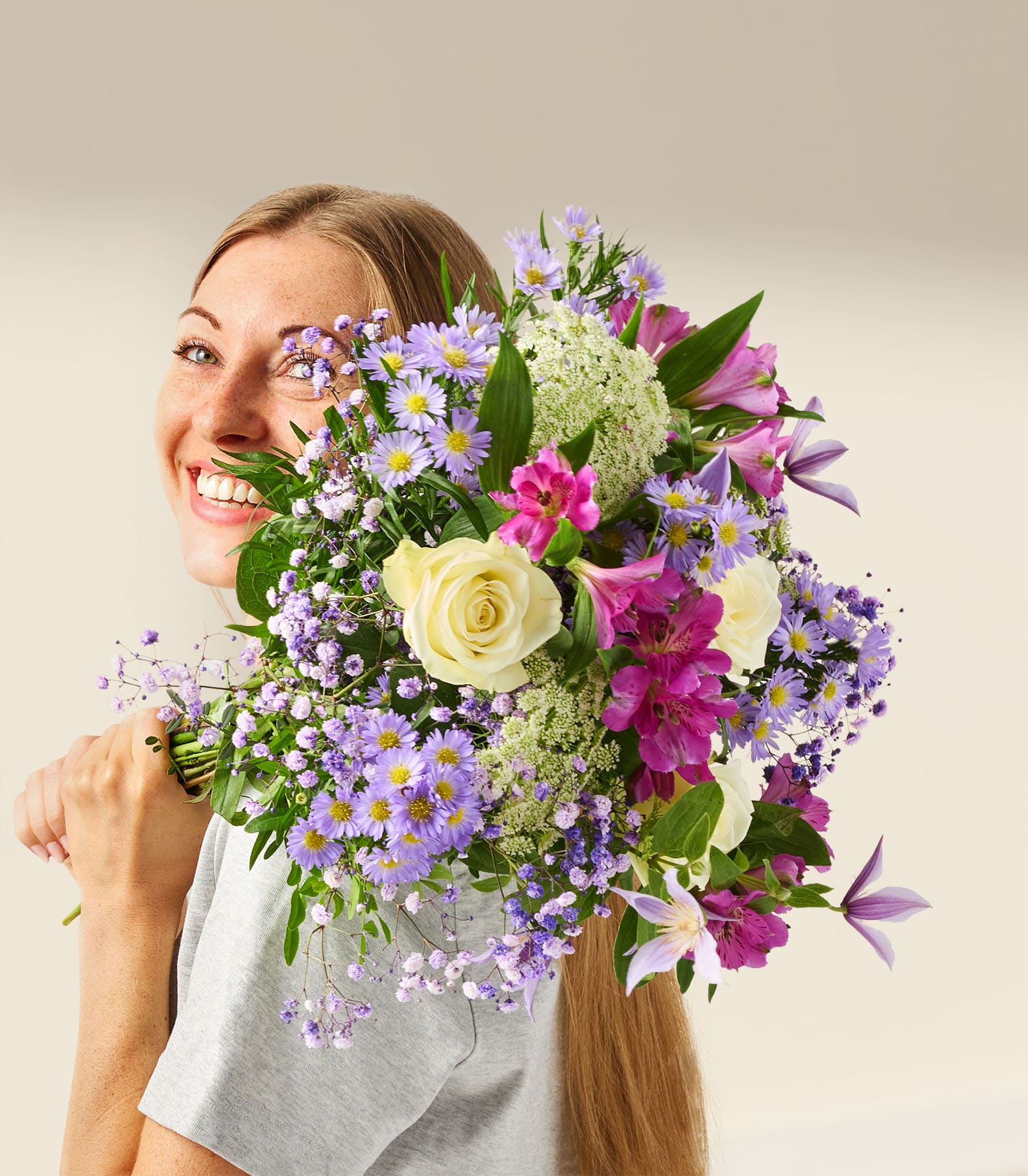 Eine lächelnde Frau hält vor einem schlichten weißen Hintergrund einen leuchtenden Strauß bunter Blumen, darunter Rosen, Gänseblümchen und andere Blüten.
