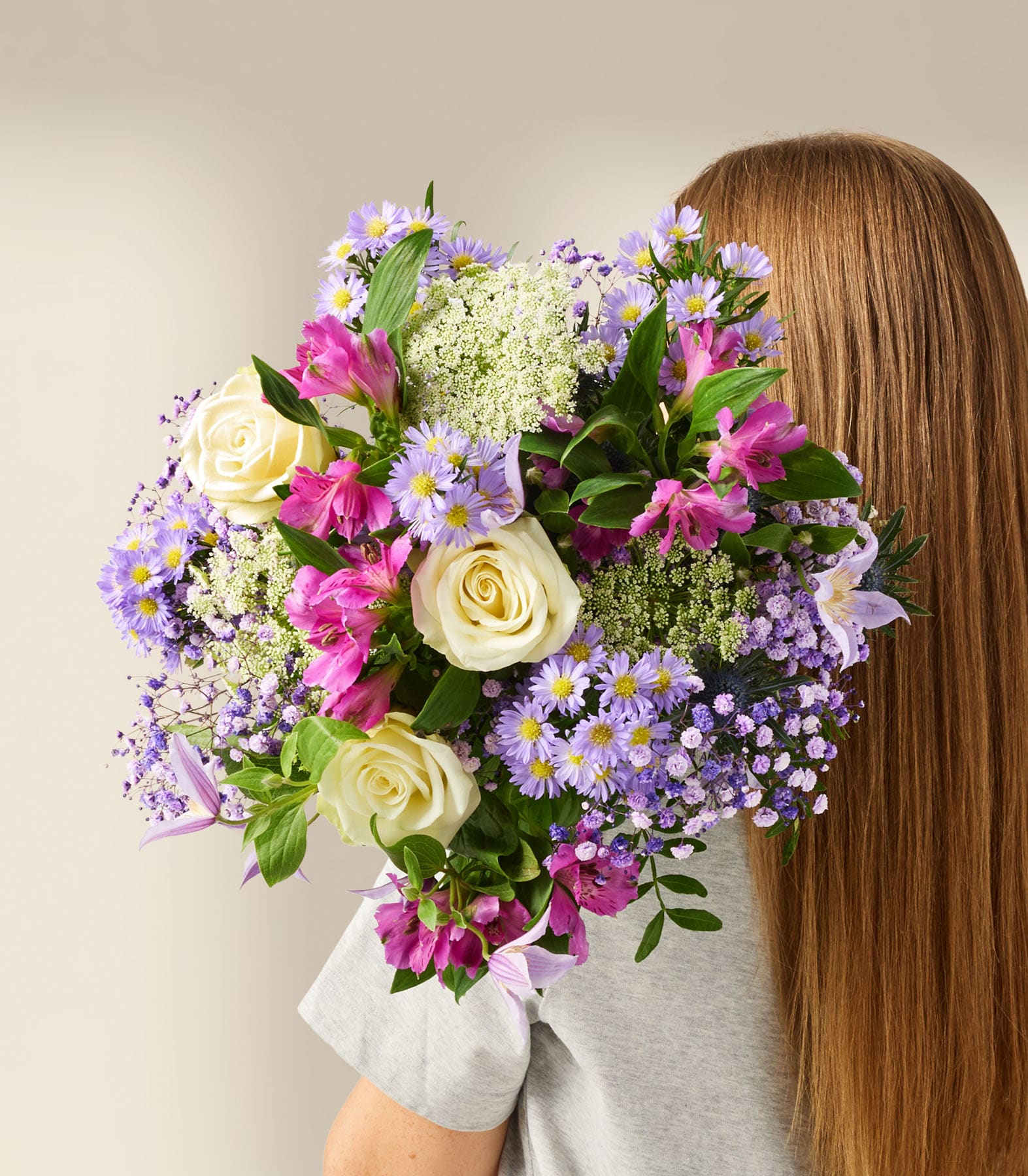 Ein farbenfroher Strauß verschiedener Blumen, darunter Rosen, Gänseblümchen und Hortensien, wird vor einem schlichten weißen Hintergrund vor eine Person mit langem braunem Haar gehalten.