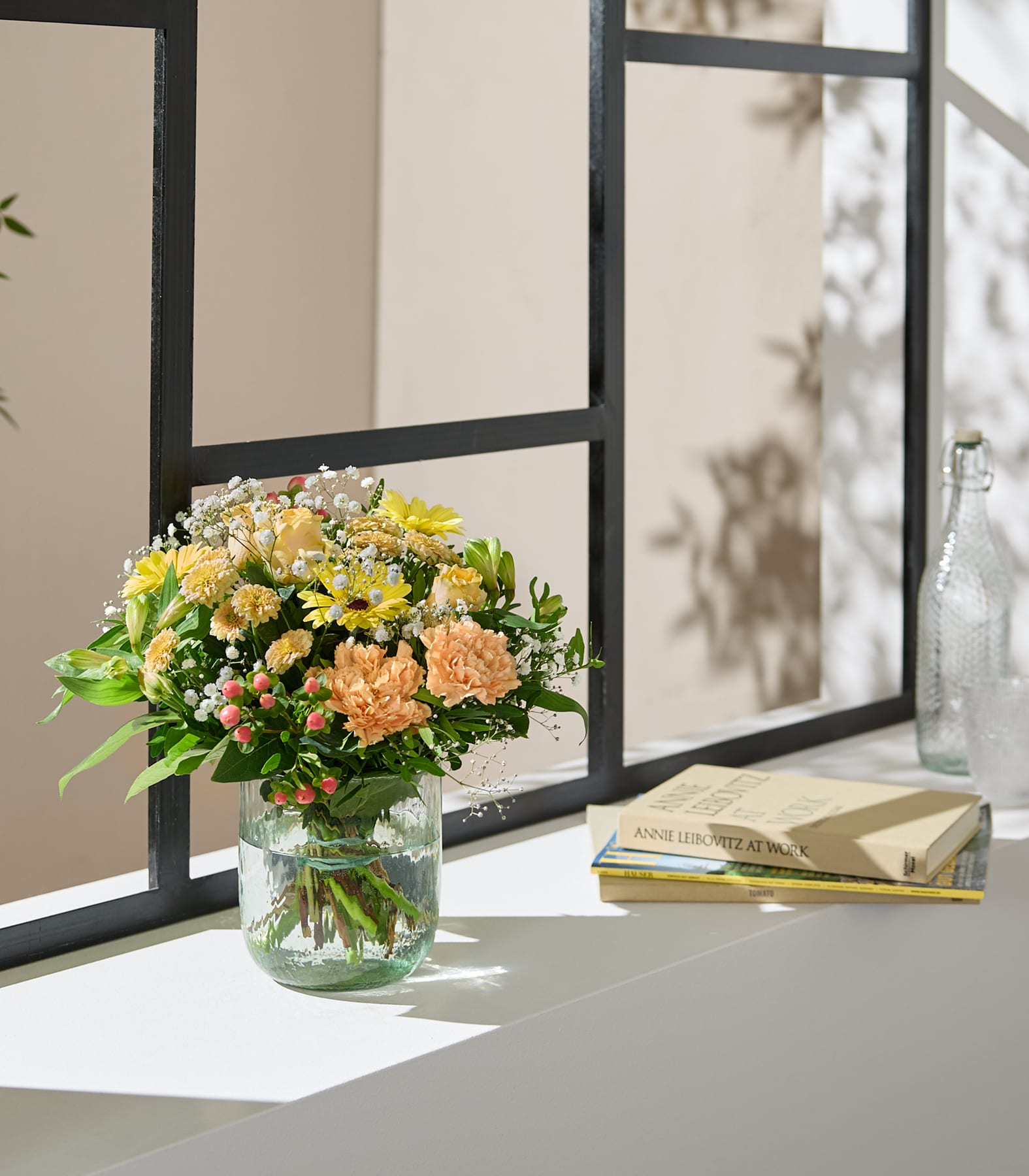 Eine Hand hält einen leuchtenden Strauß gelber und orangefarbener Blumen mit zarten weißen Füllblumen vor einem sanften, neutralen Hintergrund.