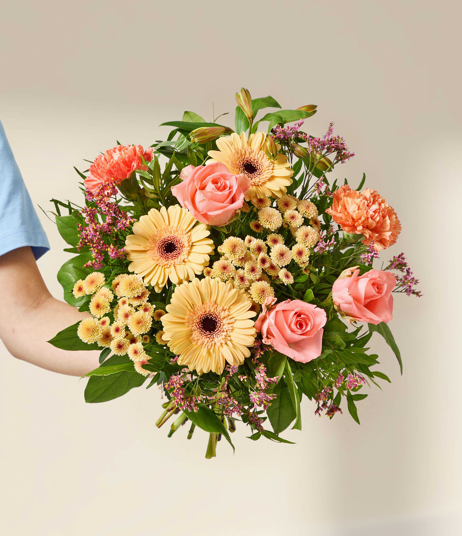 Eine Hand hält einen leuchtenden Strauß aus rosa Rosen, orangefarbenen Nelken und gelben Gerbera vor einem sanften, neutralen Hintergrund.