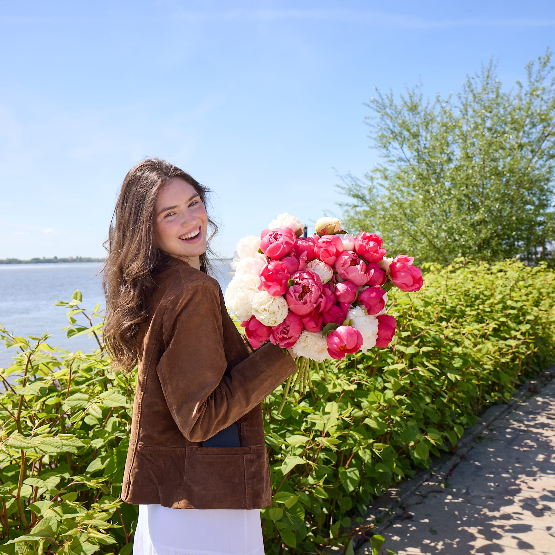 Im Vordergrund hält eine lächelnde Frau in einer braunen Jacke einen Strauß rosa und weißer Pfingstrosen; im Hintergrund sind eine üppige grüne Hecke und ein Gewässer unter klarem blauen Himmel zu sehen.