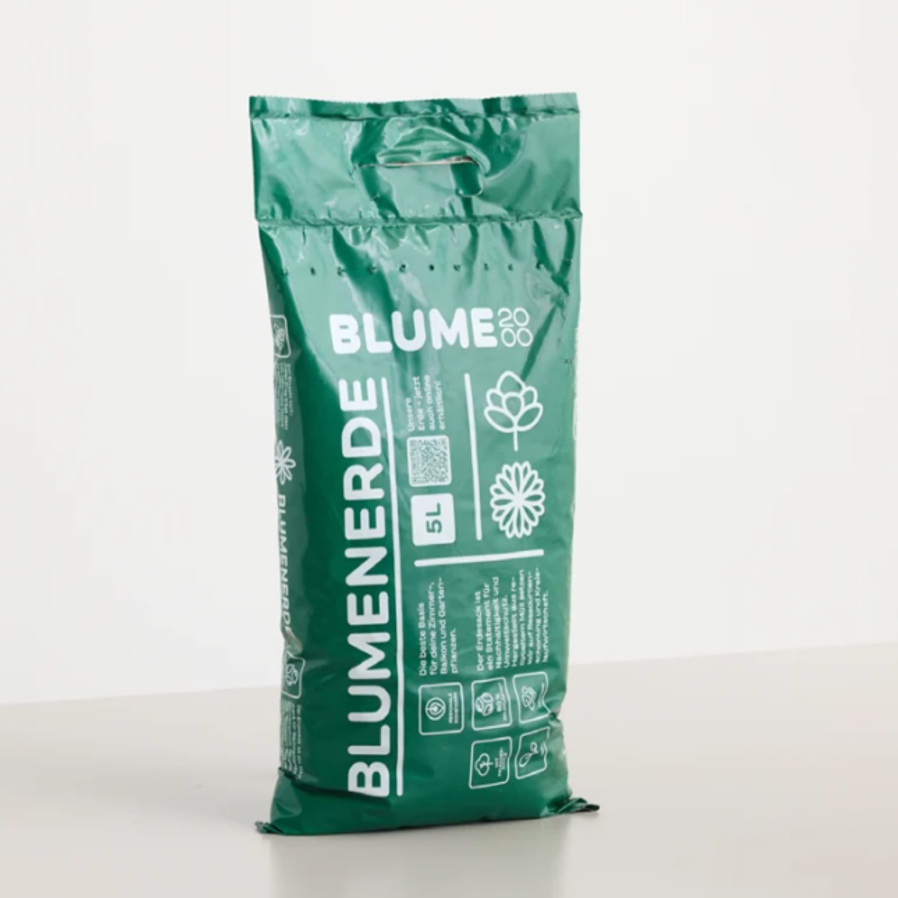 Ein grüner Sack Blumenerde mit dem Markennamen "BLUME" ist auf einer hellgrauen Fläche vor einem schlichten weißen Hintergrund zentriert.