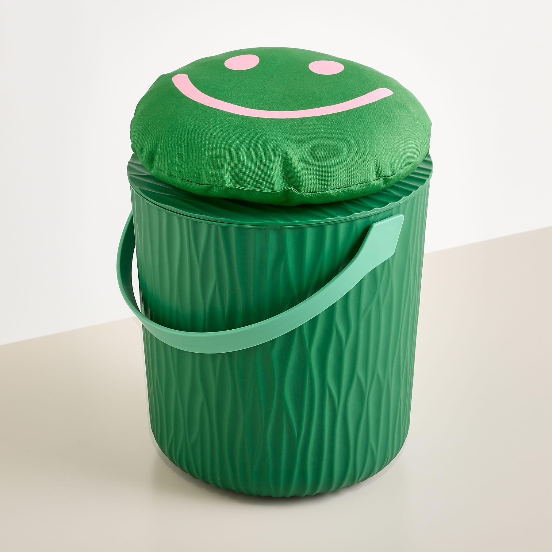 Ein grüner Eimer mit einem Kissen in Form eines Smileys darauf steht auf einer hellen Oberfläche vor einem schlichten weißen Hintergrund.
