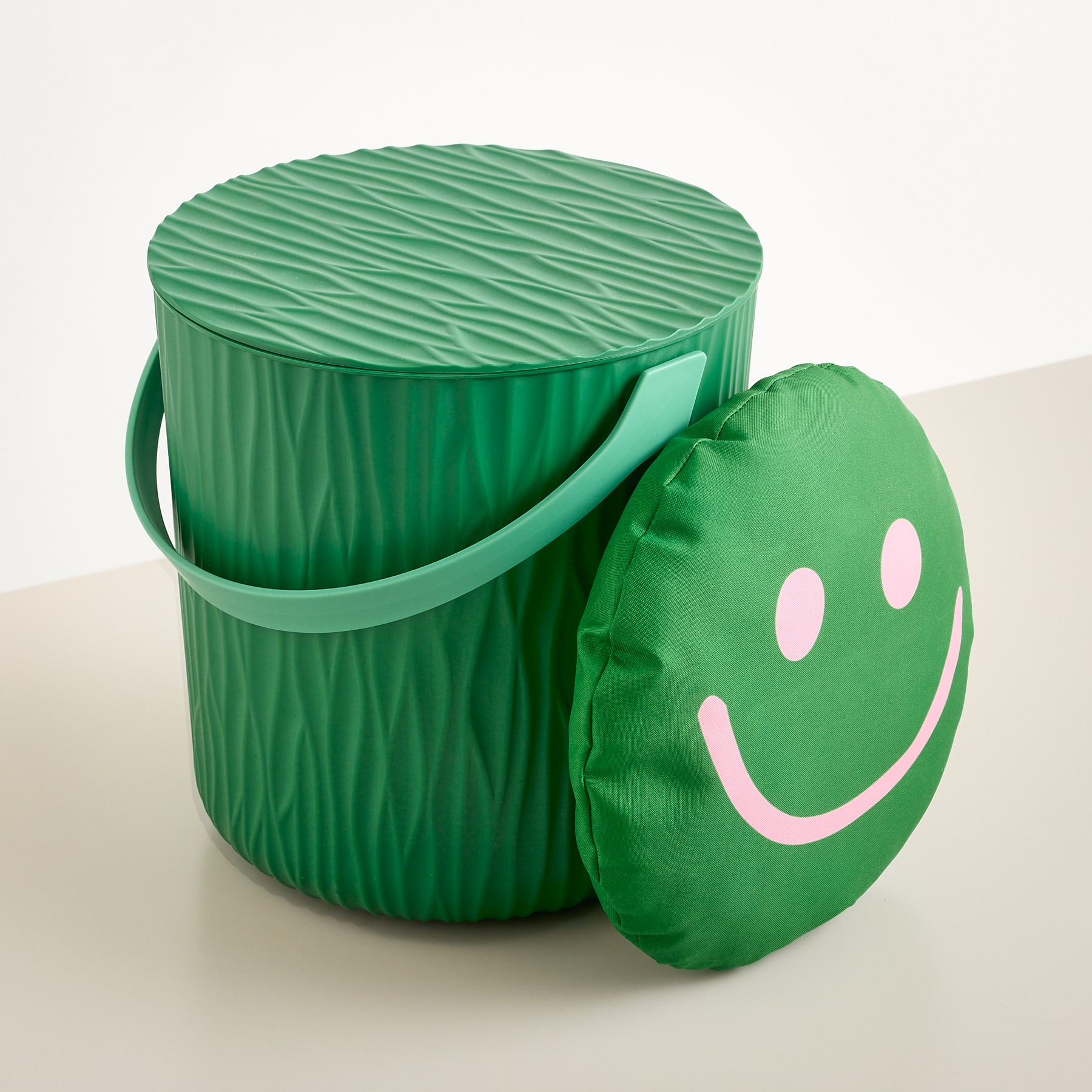 Neben einem runden grünen Kissen mit einem hellen Untergrund steht ein strukturierter grüner Eimer mit Henkel, auf dem ein rosa Smiley-Gesicht abgebildet ist.