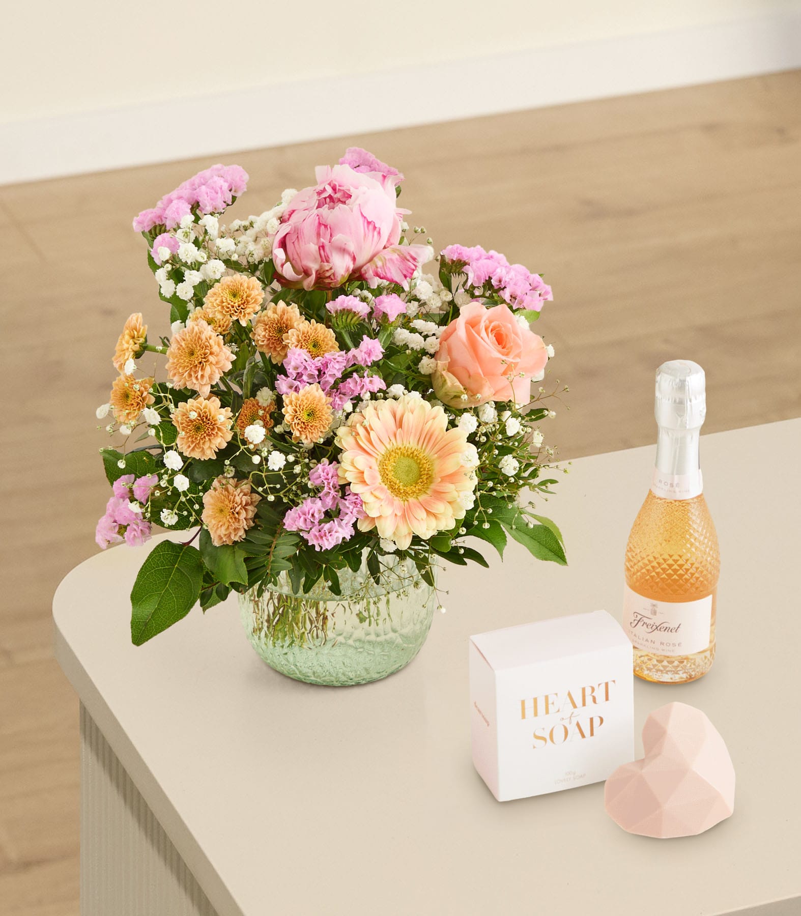 Ein leuchtender Strauß rosa und orangefarbener Blumen steht in einer Glasvase auf einem hellen Tisch. Im Vordergrund befinden sich eine kleine Flasche Champagner, eine Seifenschachtel und ein herzförmiger Gegenstand. Das Ganze ist vor einem sanft beleuchteten, neutralen Hintergrund angeordnet.