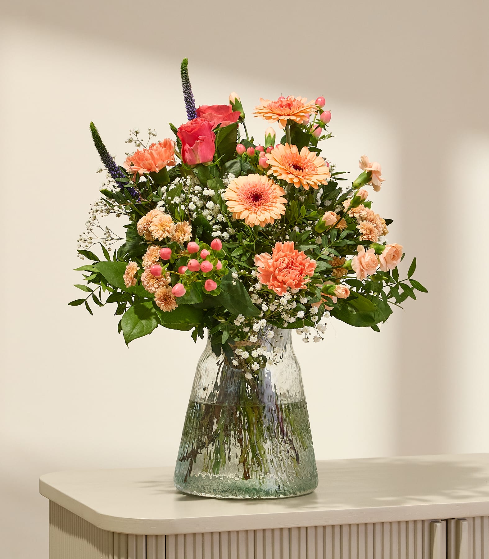 Ein leuchtender Strauß rosa und orangefarbener Blumen füllt eine strukturierte Glasvase auf einer hellen Oberfläche, im Hintergrund wirft eine sanft beleuchtete Wand Schatten. (Beschreibungstext KI-generiert)