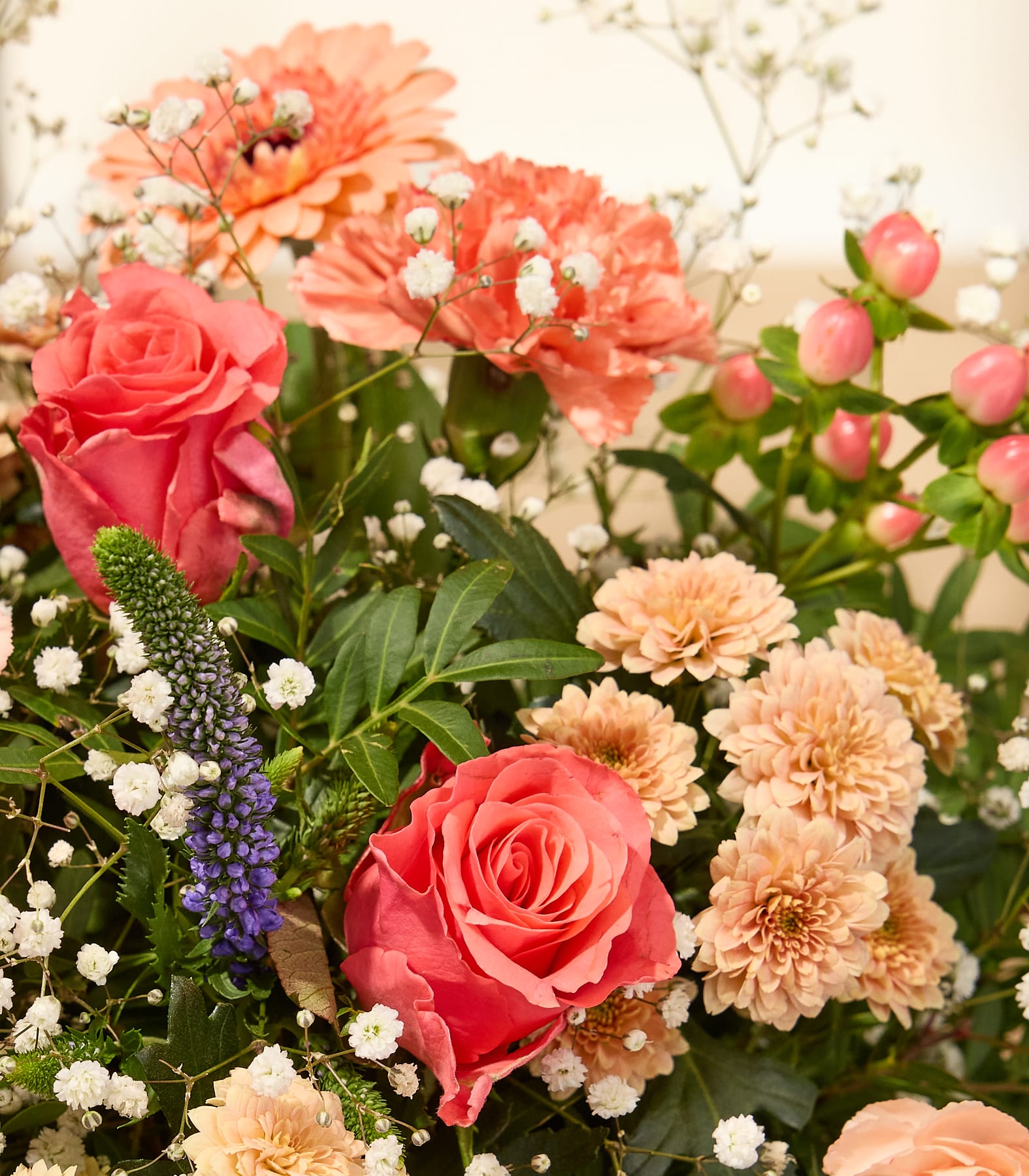 Eine junge Frau mit langem, dunklem Haar hält vor einem schlichten weißen Hintergrund einen großen, leuchtenden Strauß bunter Blumen, darunter Gerbera, Rosen und Grünpflanzen. (Beschreibungstext KI-generiert)