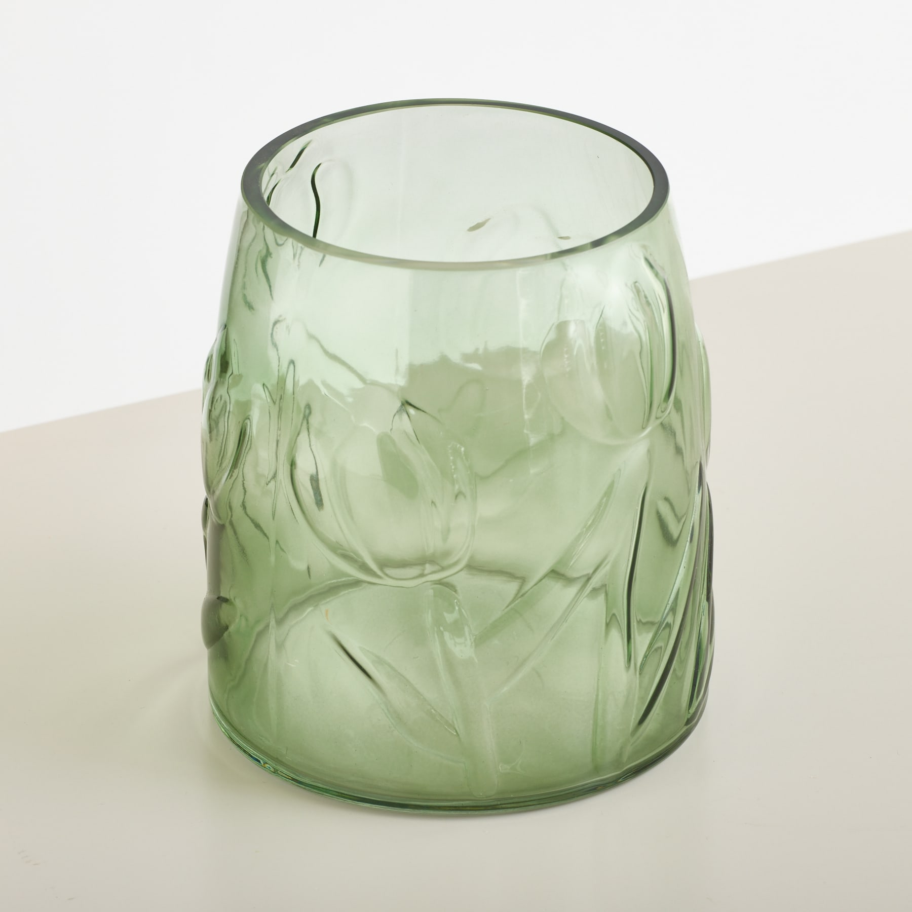 Eine durchscheinende grüne Glasvase mit erhabenem Blumenmuster steht auf einer weißen Fläche vor einem schlichten weißen Hintergrund.