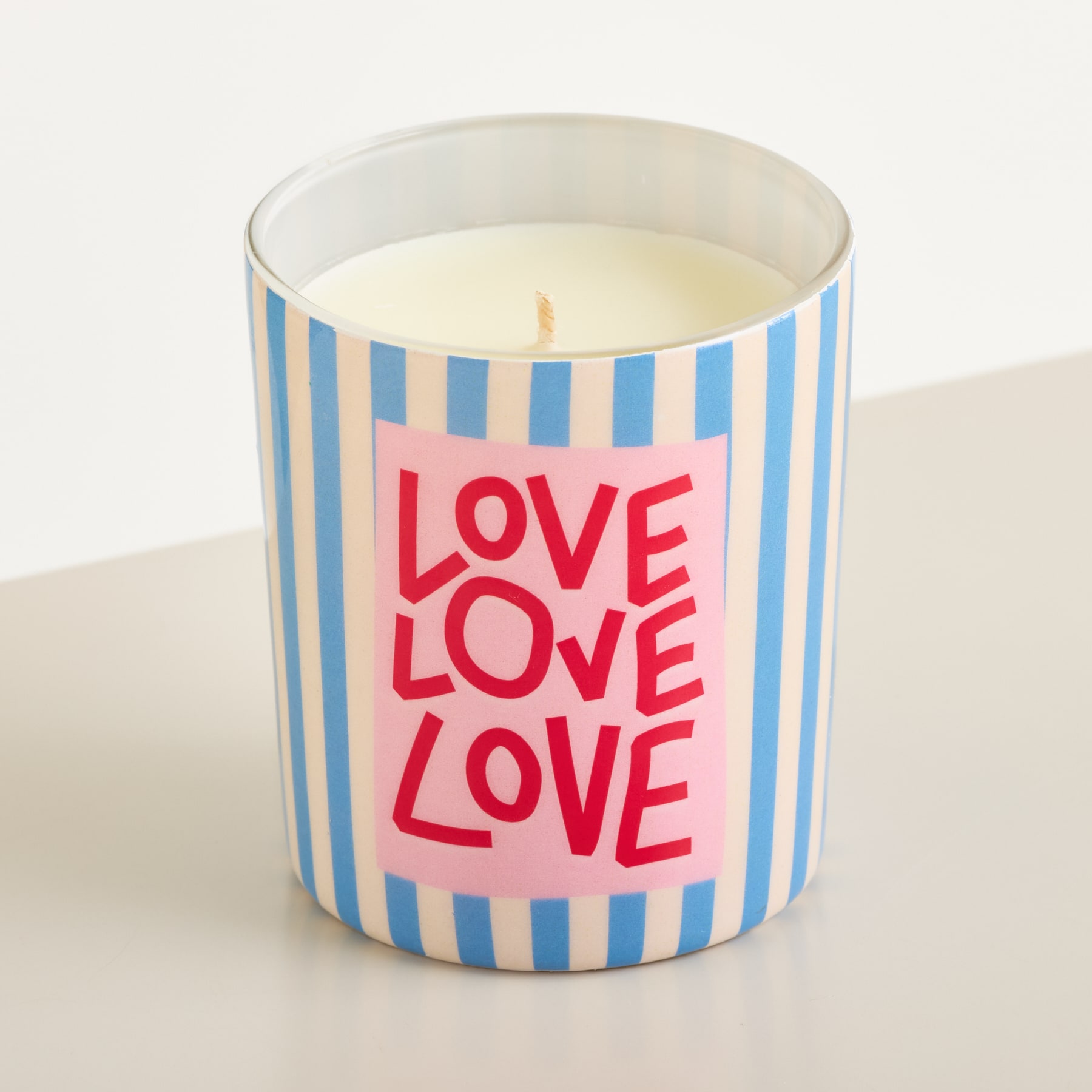 Eine gestreifte Kerze mit der Aufschrift „LOVE LOVE LOVE“ steht auf einer weißen Fläche vor einem schlichten weißen Hintergrund.