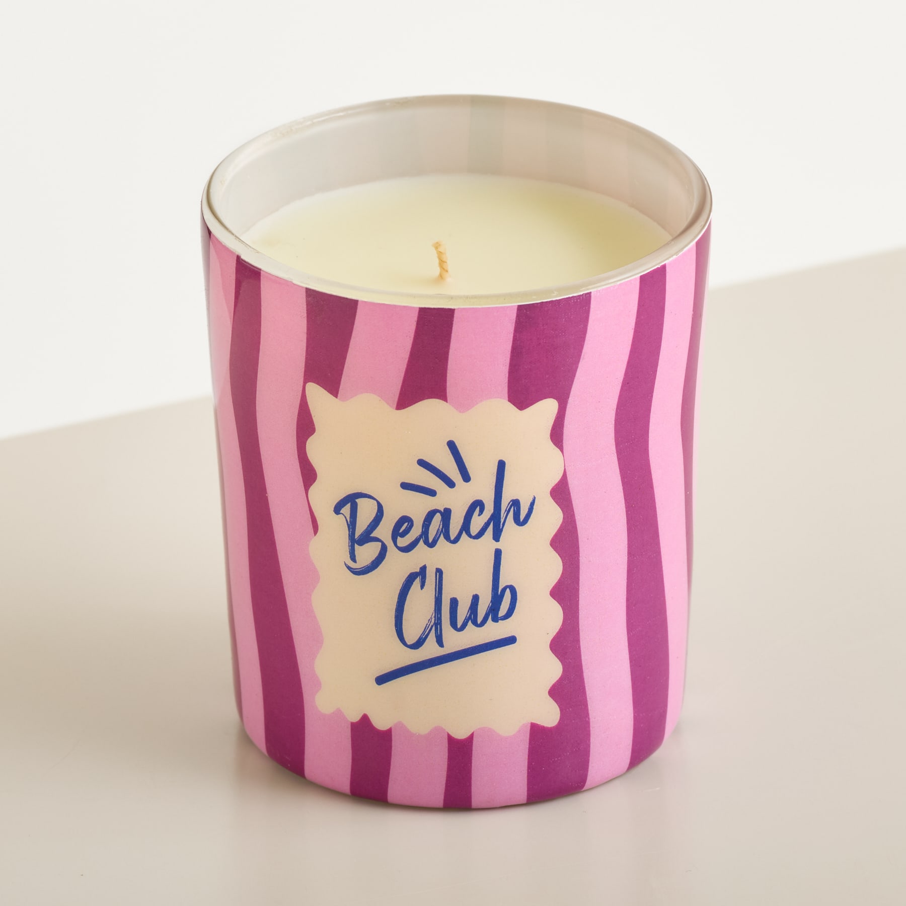 Eine rosa-lila gestreifte Kerze mit der Aufschrift „Beach Club“ steht auf einer weißen Oberfläche mit hellgrauem Hintergrund.