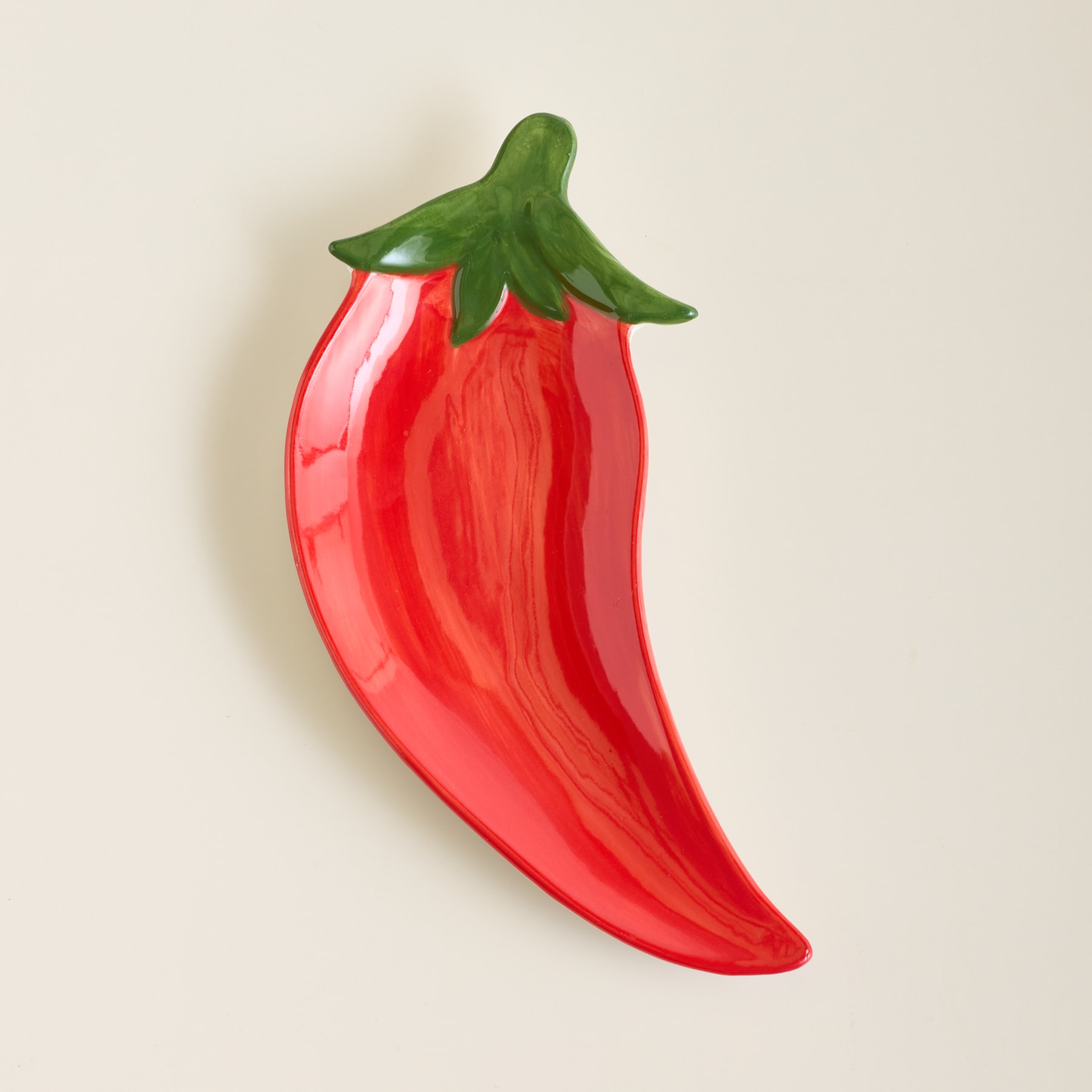 Eine glänzende, rote, chili-förmige Schale mit grünem Stiel steht auf einem schlichten, hellen Hintergrund.