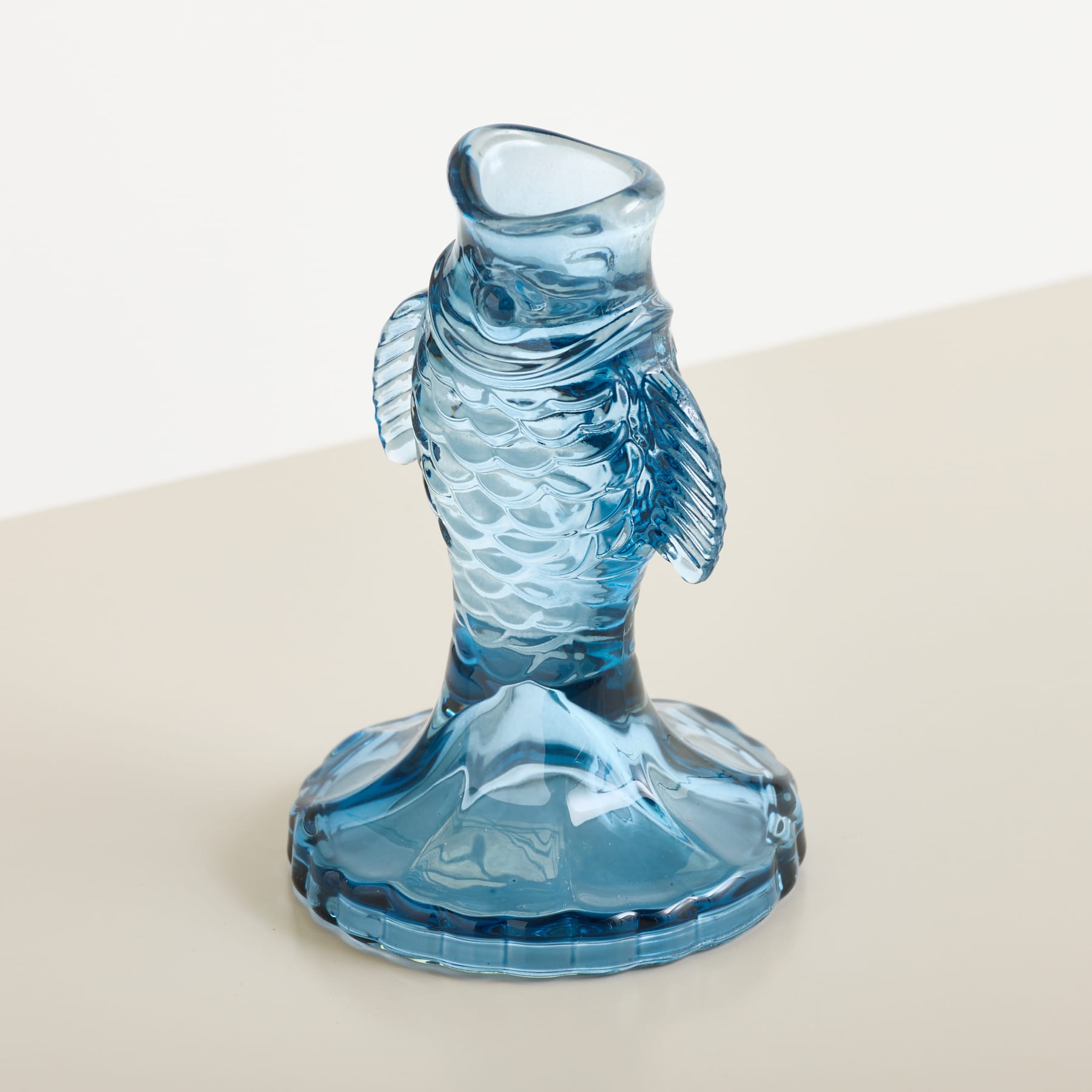 Eine blaue, fischförmige Glasvase mit aufwendigen Schuppendetails steht auf einer hellen Oberfläche vor einem schlichten, cremeweißen Hintergrund.
