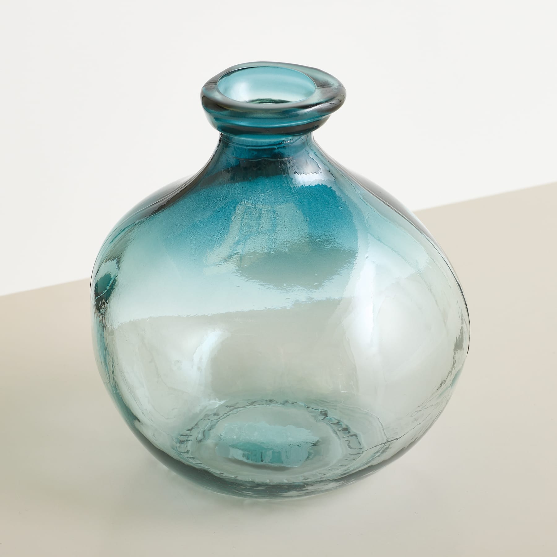 Eine durchsichtige Glasvase mit blauem Farbverlauf steht auf einer hellen Oberfläche vor einem sanft beleuchteten, neutralen Hintergrund.
