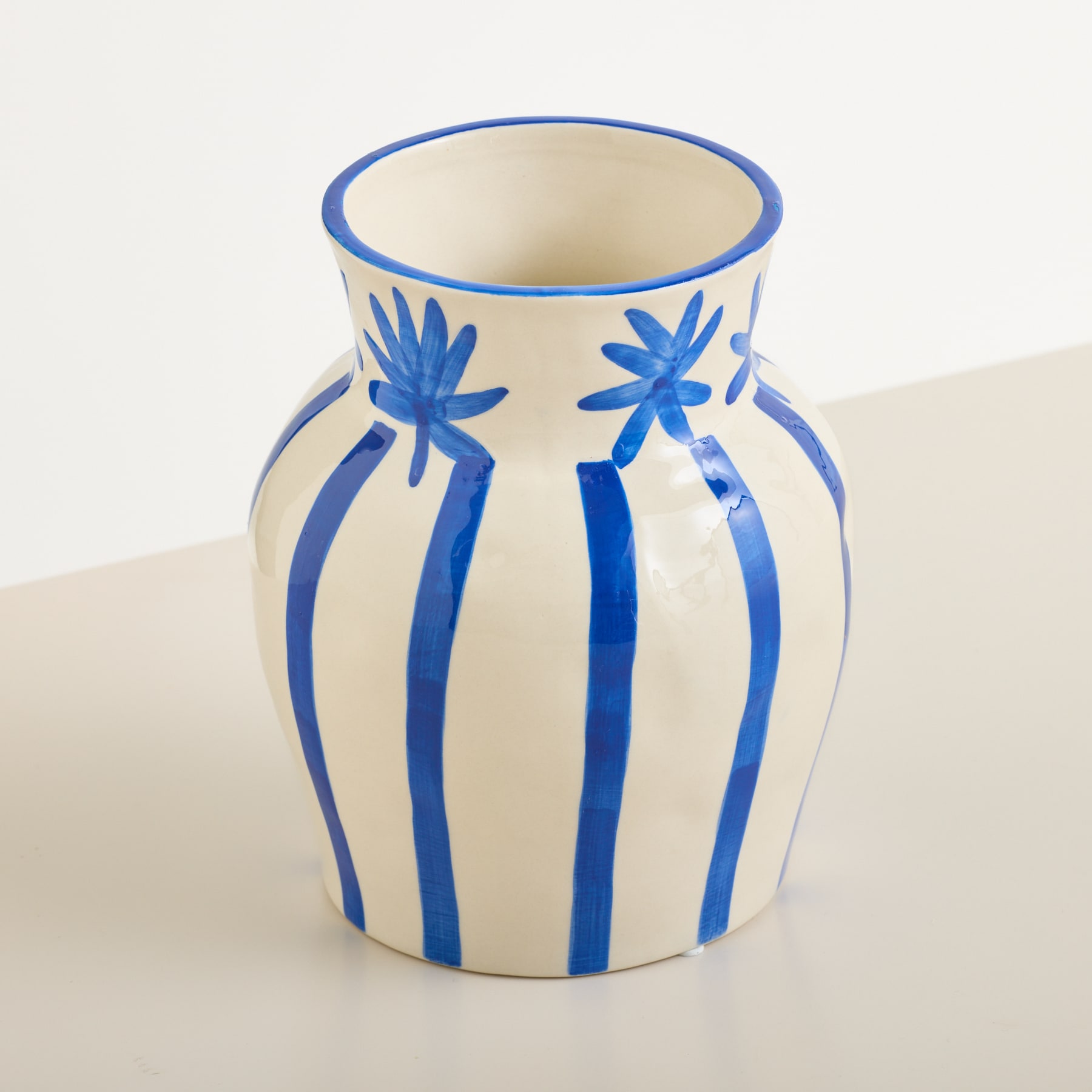Eine cremefarbene Vase mit blauen Streifen und floralen Verzierungen steht auf einer weißen Fläche vor einem schlichten weißen Hintergrund.