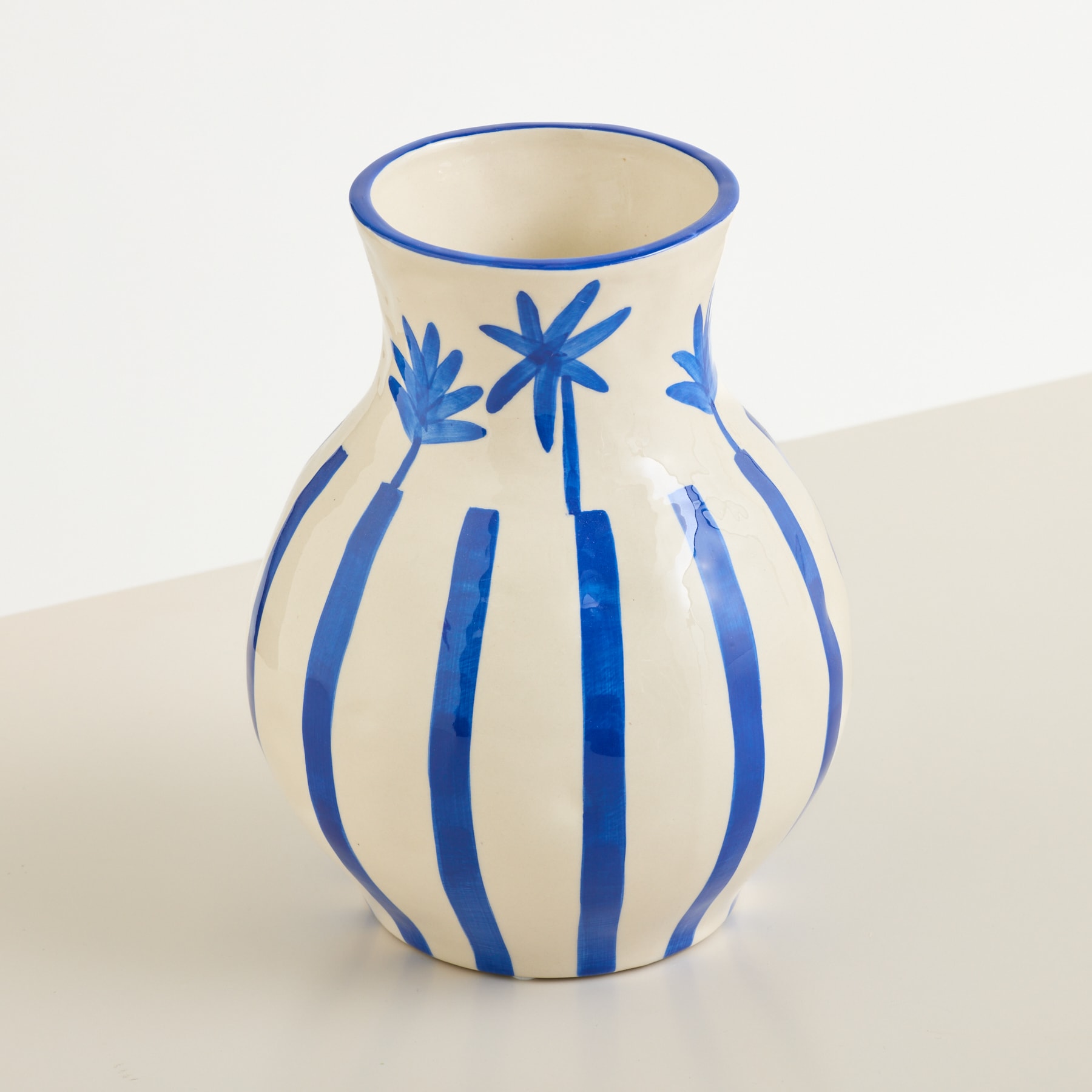 Eine cremefarbene Vase mit blauen Streifen und Palmenblattmotiven steht auf einer weißen Fläche vor einem schlichten weißen Hintergrund.