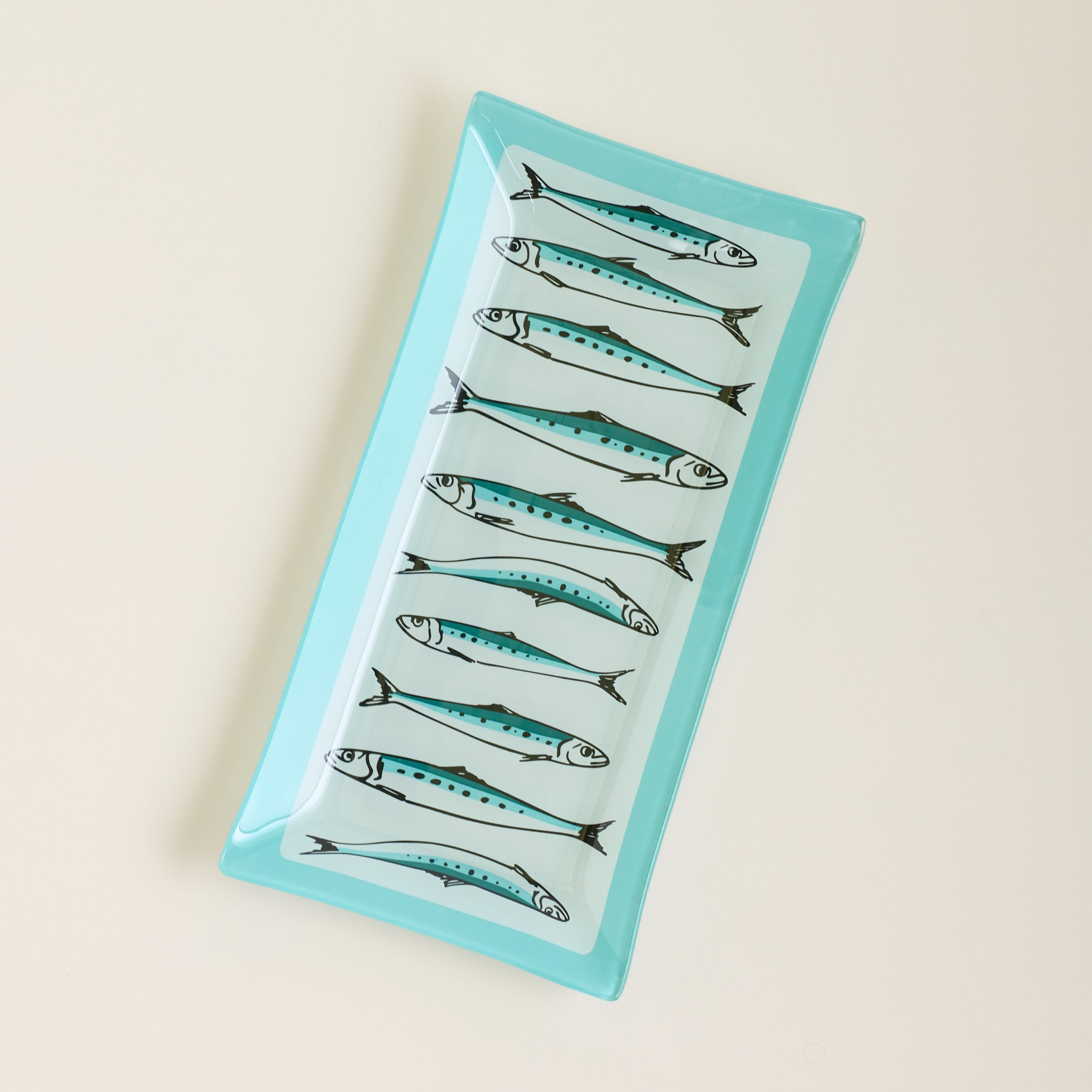 Ein rechteckiges Glastablett mit einem Muster aus blauen Fischen steht vor einem schlichten, hellen Hintergrund.