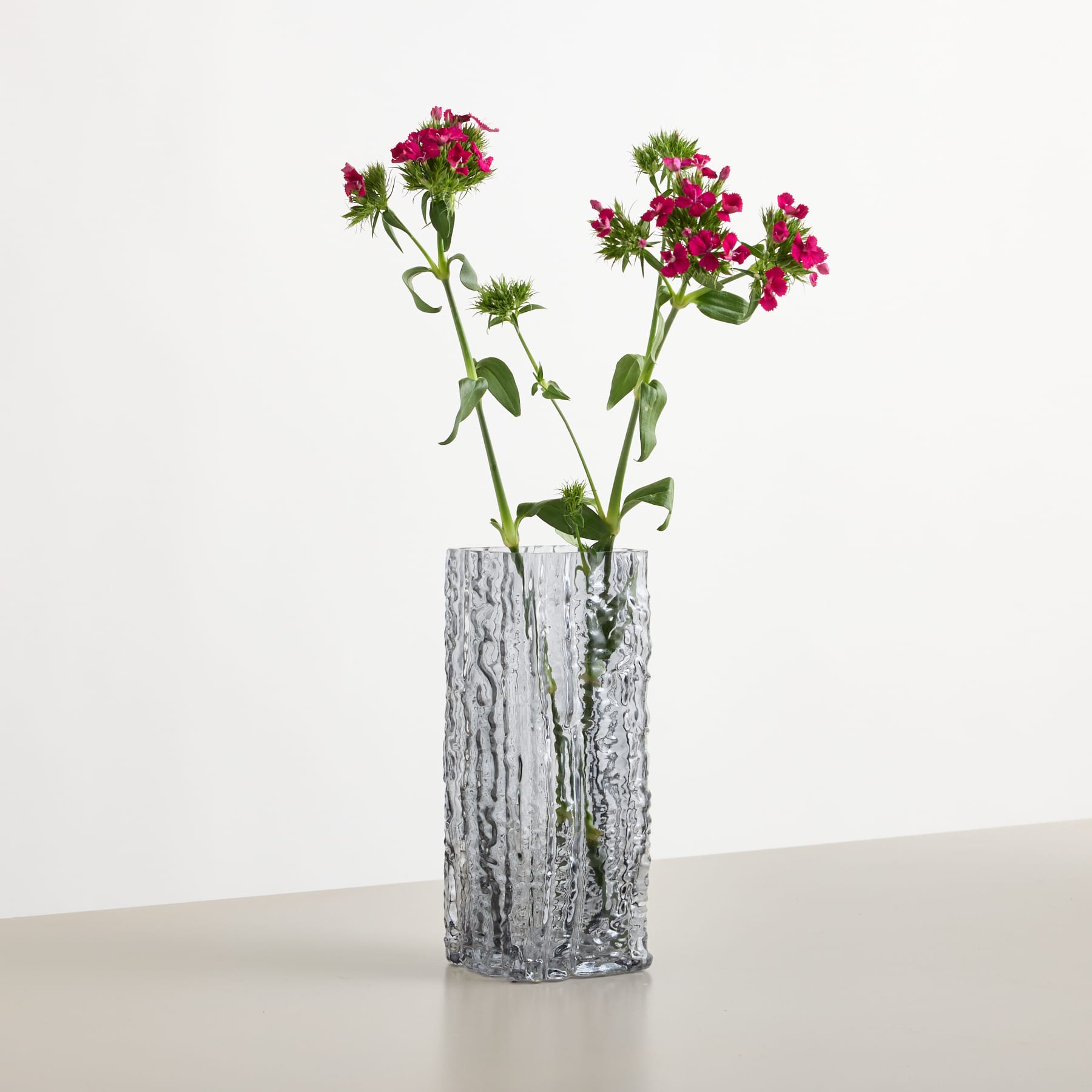 Eine strukturierte Glasvase mit rosa Blumen steht auf einer hellen Oberfläche vor einem schlichten, hellen Hintergrund.
