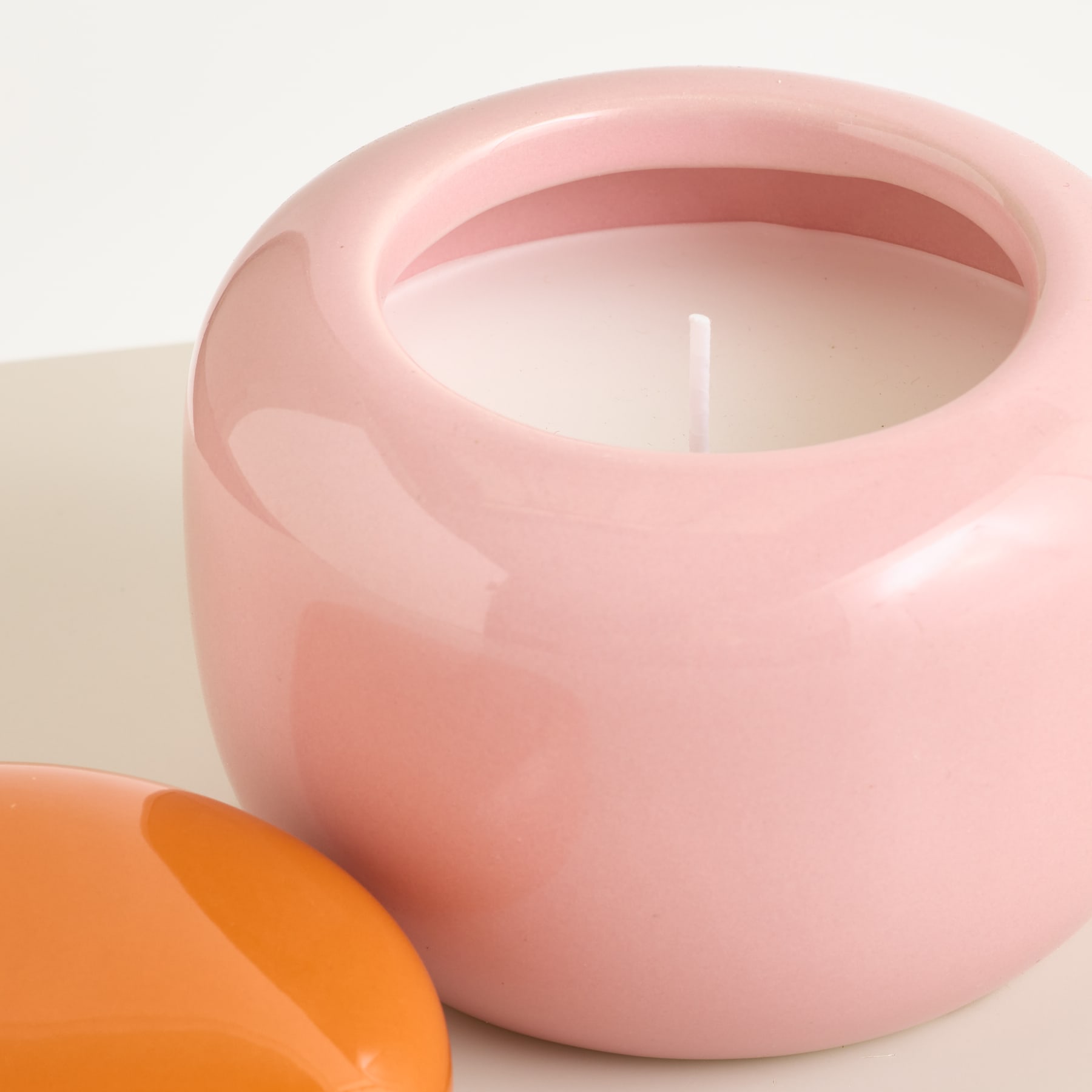 Im Vordergrund steht eine rosa Keramikkerze neben einem orangefarbenen Gegenstand; der Hintergrund ist schlicht.