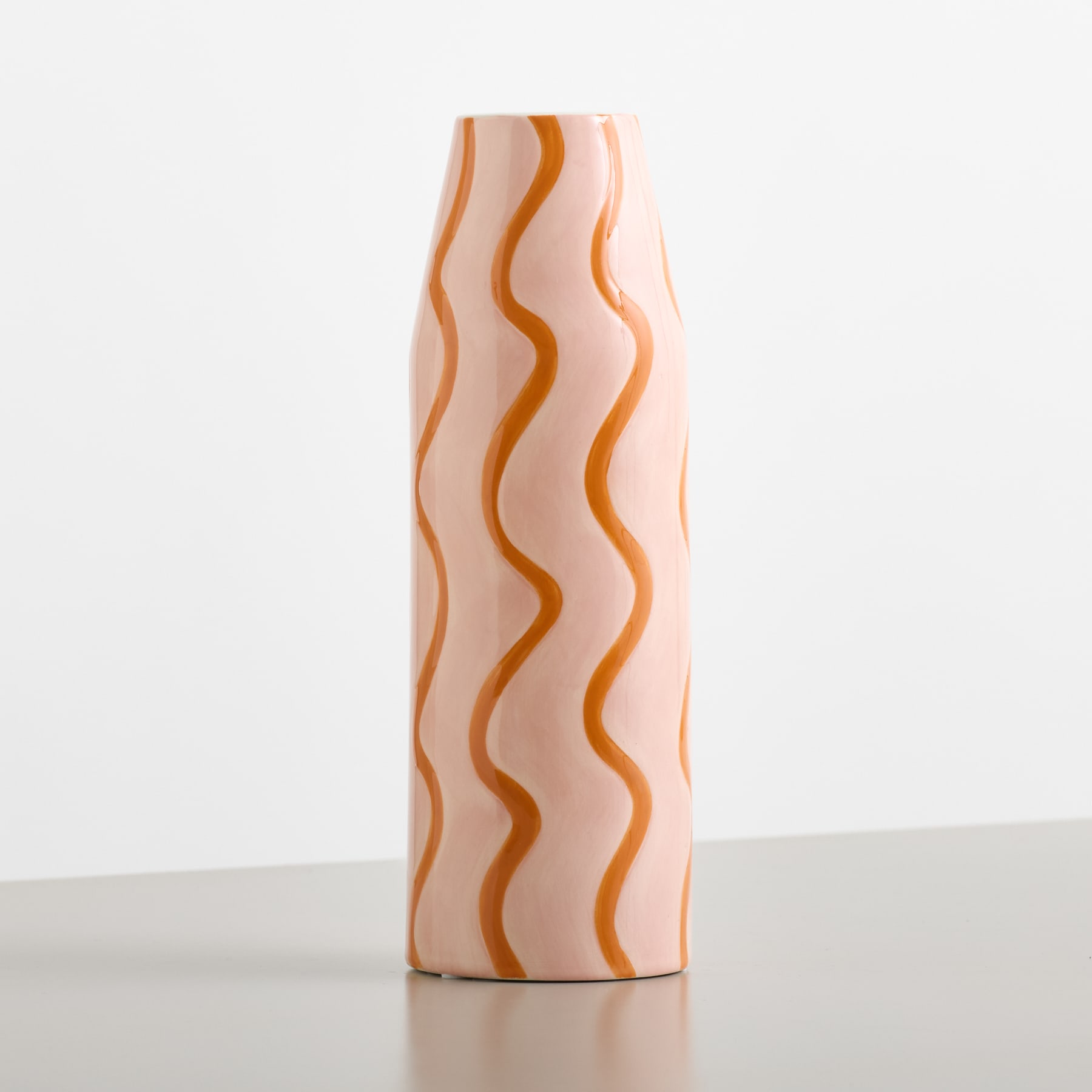 Eine hohe, hellrosa Vase mit wellenförmigen orangefarbenen Linien steht auf einer grauen Fläche vor einem schlichten weißen Hintergrund.