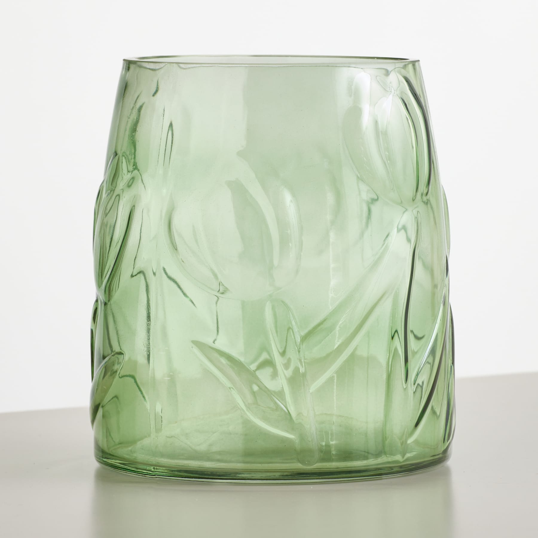 Eine grüne Glasvase mit geprägten Blumenmustern steht auf einer hellgrauen Oberfläche vor einem schlichten weißen Hintergrund.