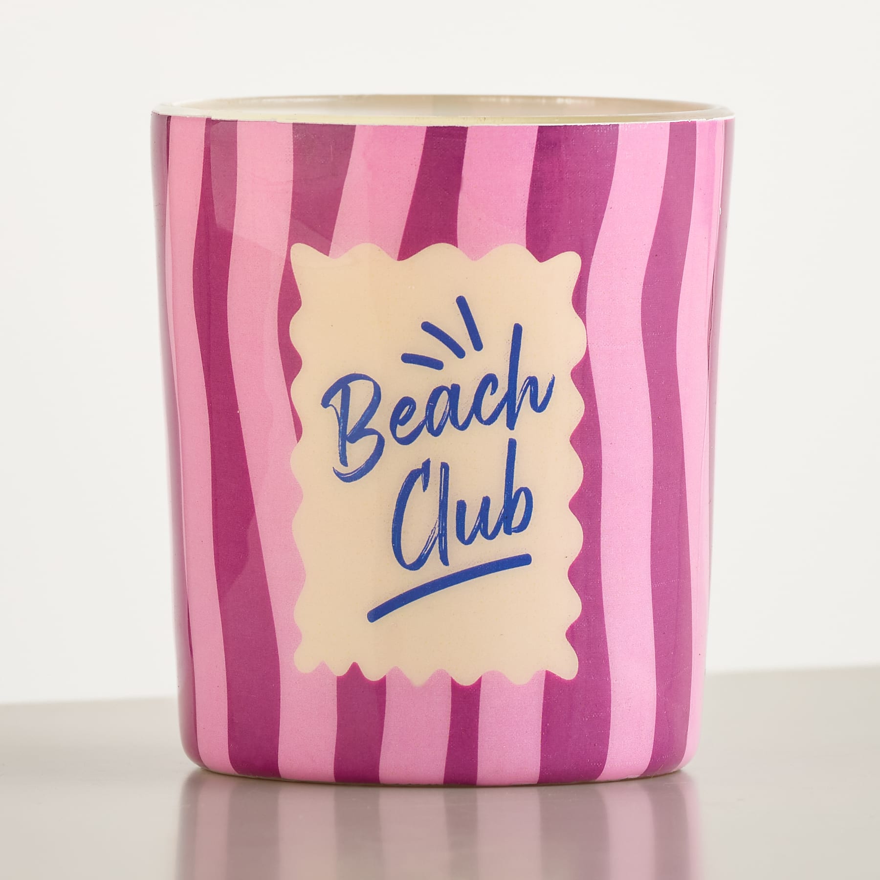 Ein rosa-lila gestreifter Becher mit der Aufschrift „Beach Club“ steht auf einer reflektierenden Oberfläche vor einem schlichten weißen Hintergrund.