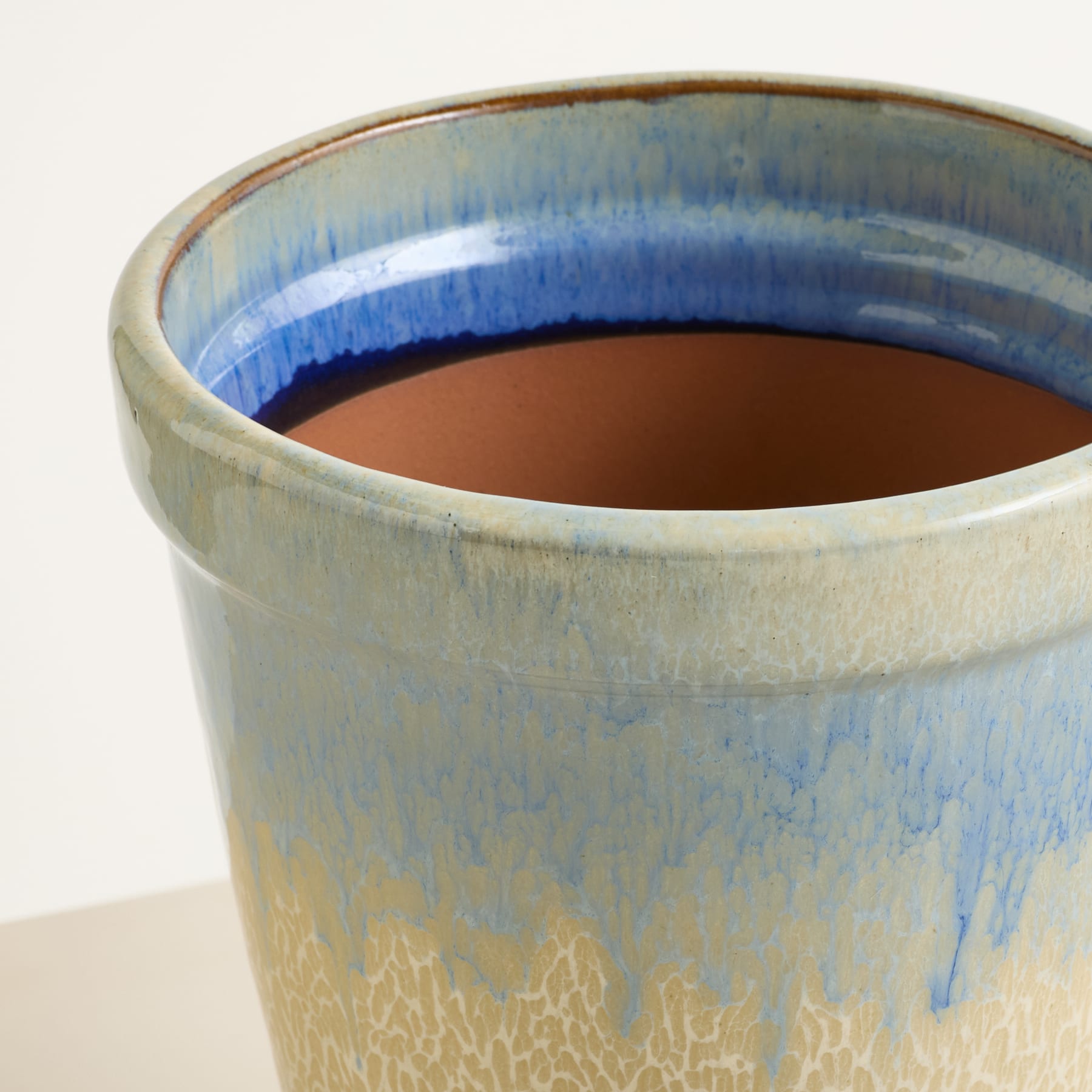 Nahaufnahme eines Keramiktopfes mit blau-brauner Glasur, gefüllt mit dunkler Erde, der auf einer hellen Oberfläche steht.