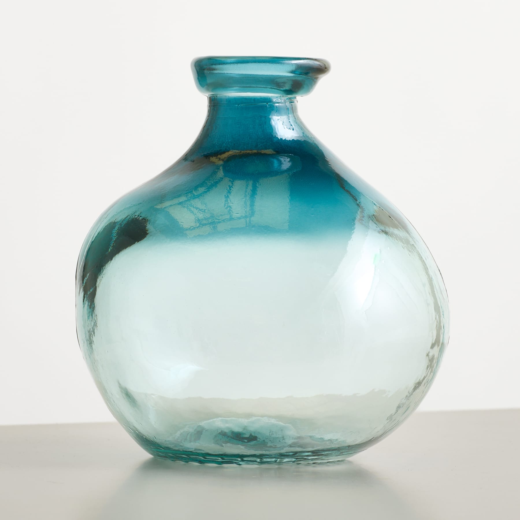 Eine runde, türkis getönte Glasvase mit glatter, reflektierender Oberfläche steht auf einer grauen Fläche vor einem schlichten weißen Hintergrund.