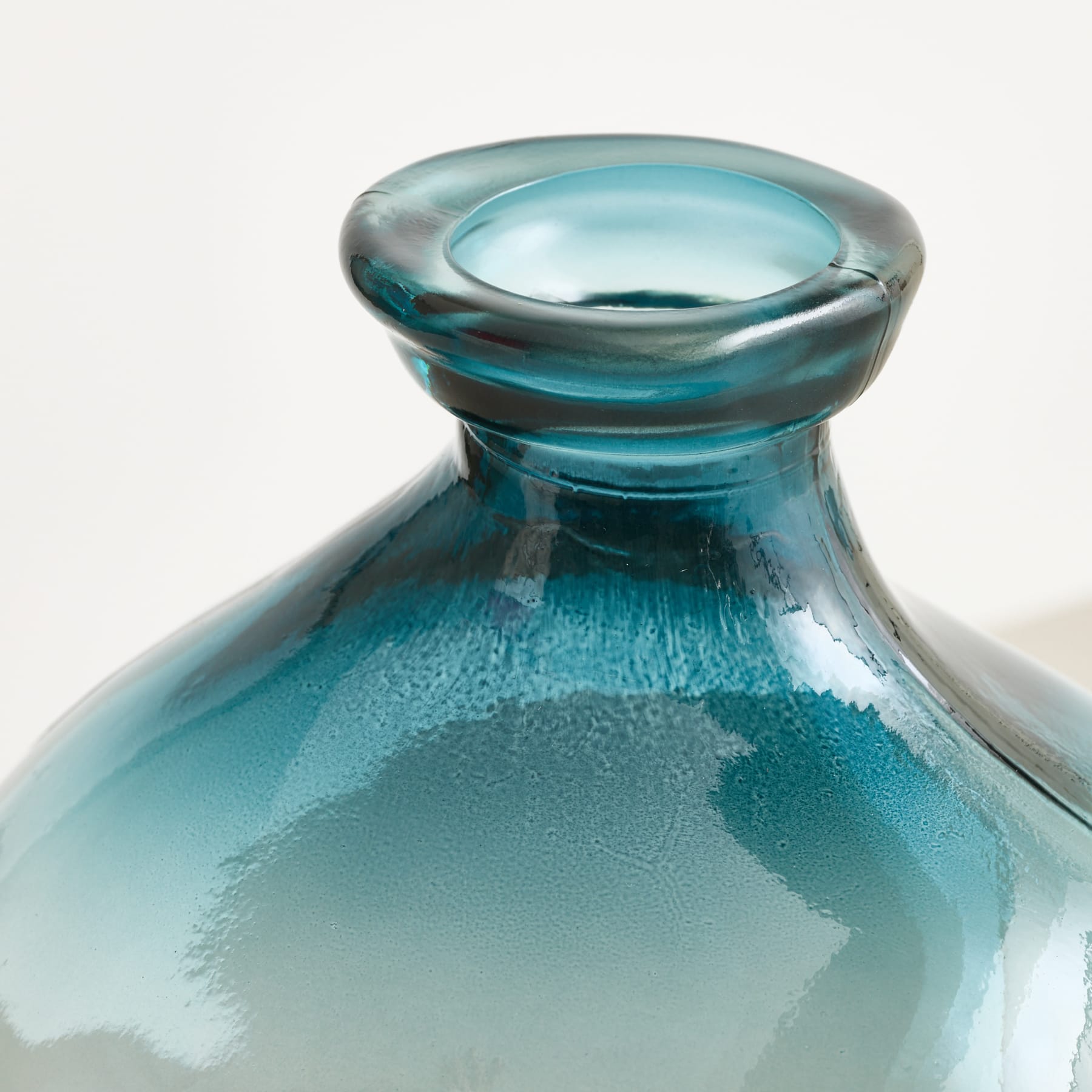 Eine Nahaufnahme des Halses und der Schulter einer türkisfarbenen Glasflasche geht in einen weichen, hellen Hintergrund über.