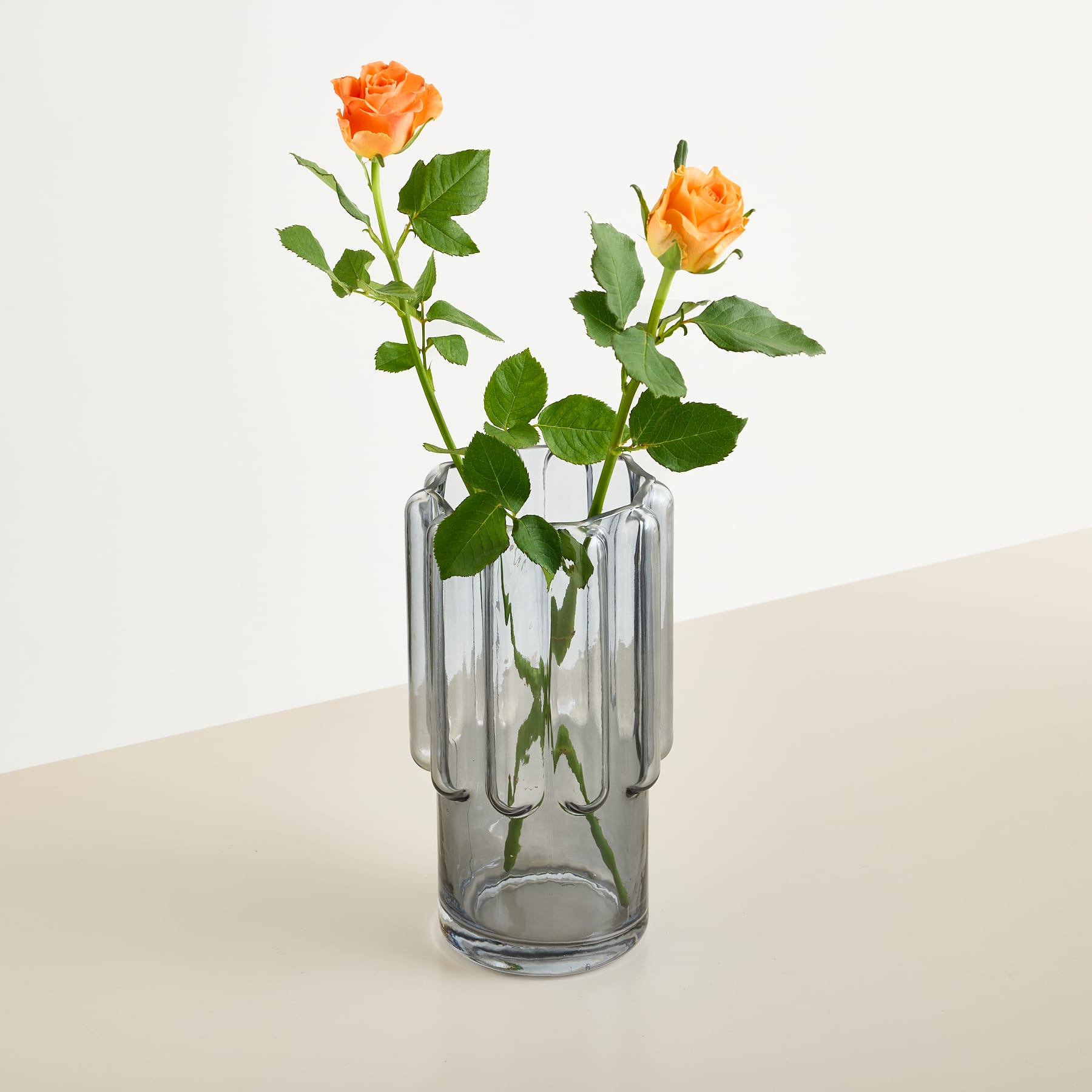 Zwei orangefarbene Rosen mit grünen Blättern stehen in einer gerippten grauen Glasvase vor einem schlichten, hellen Hintergrund.