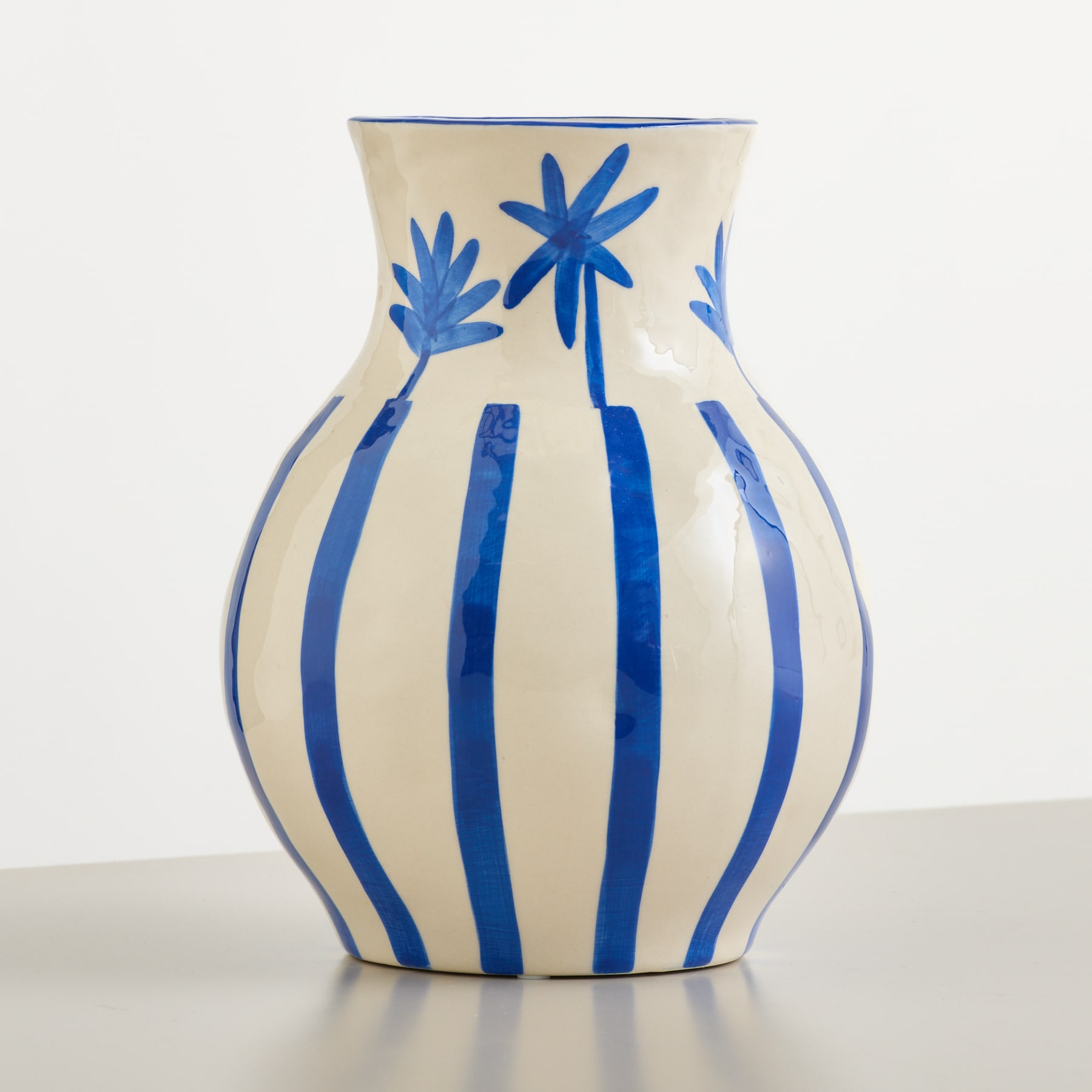 Eine cremefarbene Vase mit blauen Streifen und Palmenblattmotiven steht auf einer grauen Oberfläche vor weißem Hintergrund.