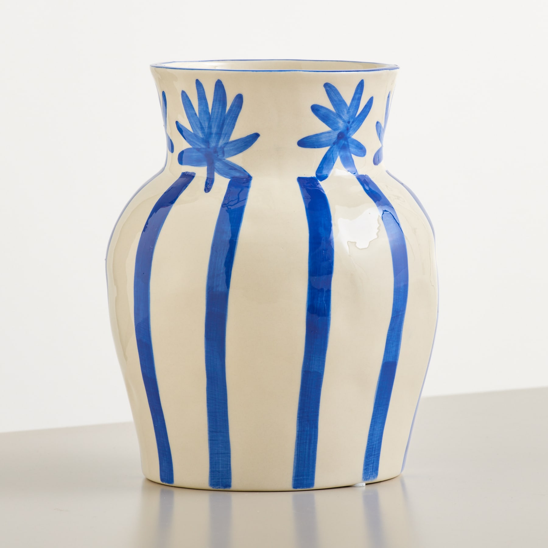 Eine cremefarbene Vase mit kräftigen blauen Streifen und floralen Verzierungen steht auf einer reflektierenden Oberfläche vor einem schlichten weißen Hintergrund.