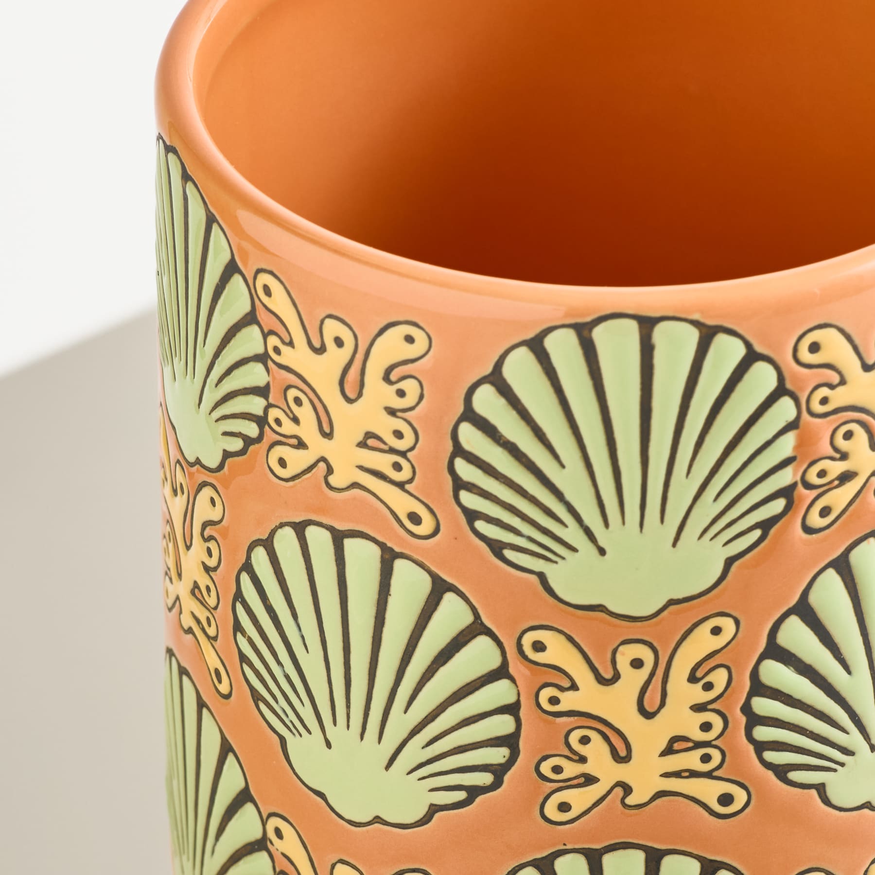 Nahaufnahme eines orangefarbenen Keramik-Pflanzgefäßes mit einem sich wiederholenden Muster aus grünen Muscheln und gelben Korallen vor einem schlichten weißen Hintergrund.
