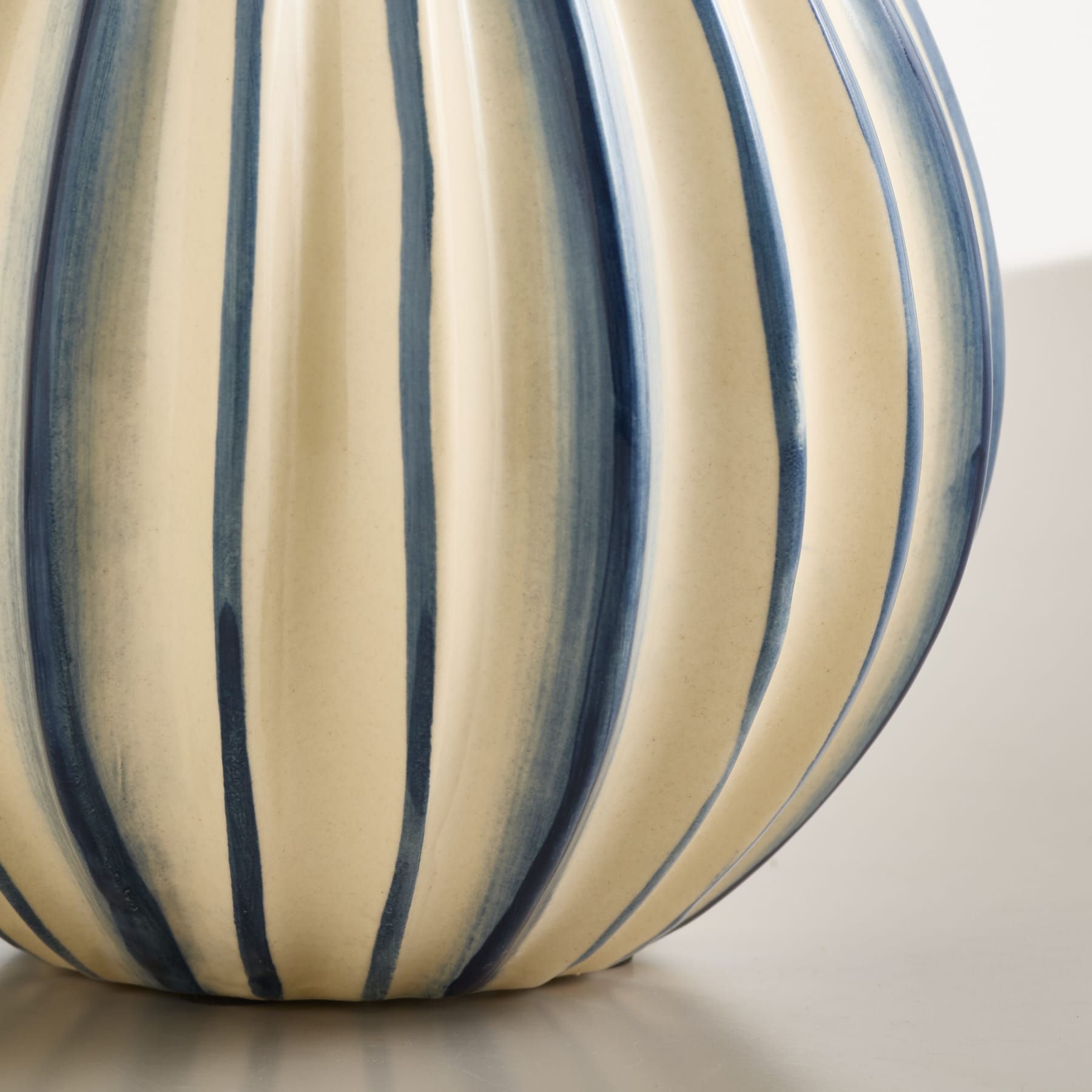 Eine cremefarbene Vase mit vertikalen blauen Streifen steht auf einer hellgrauen Fläche vor weißem Hintergrund.
