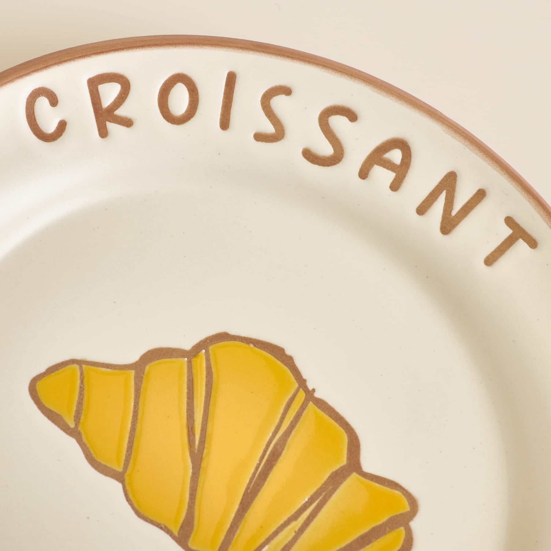 Auf einem cremefarbenen Teller mit braunem Rand ist im Vordergrund eine gelbe Croissant-Abbildung zu sehen, im Hintergrund ist das Wort „CROISSANT“ in braunen Buchstaben eingeprägt.