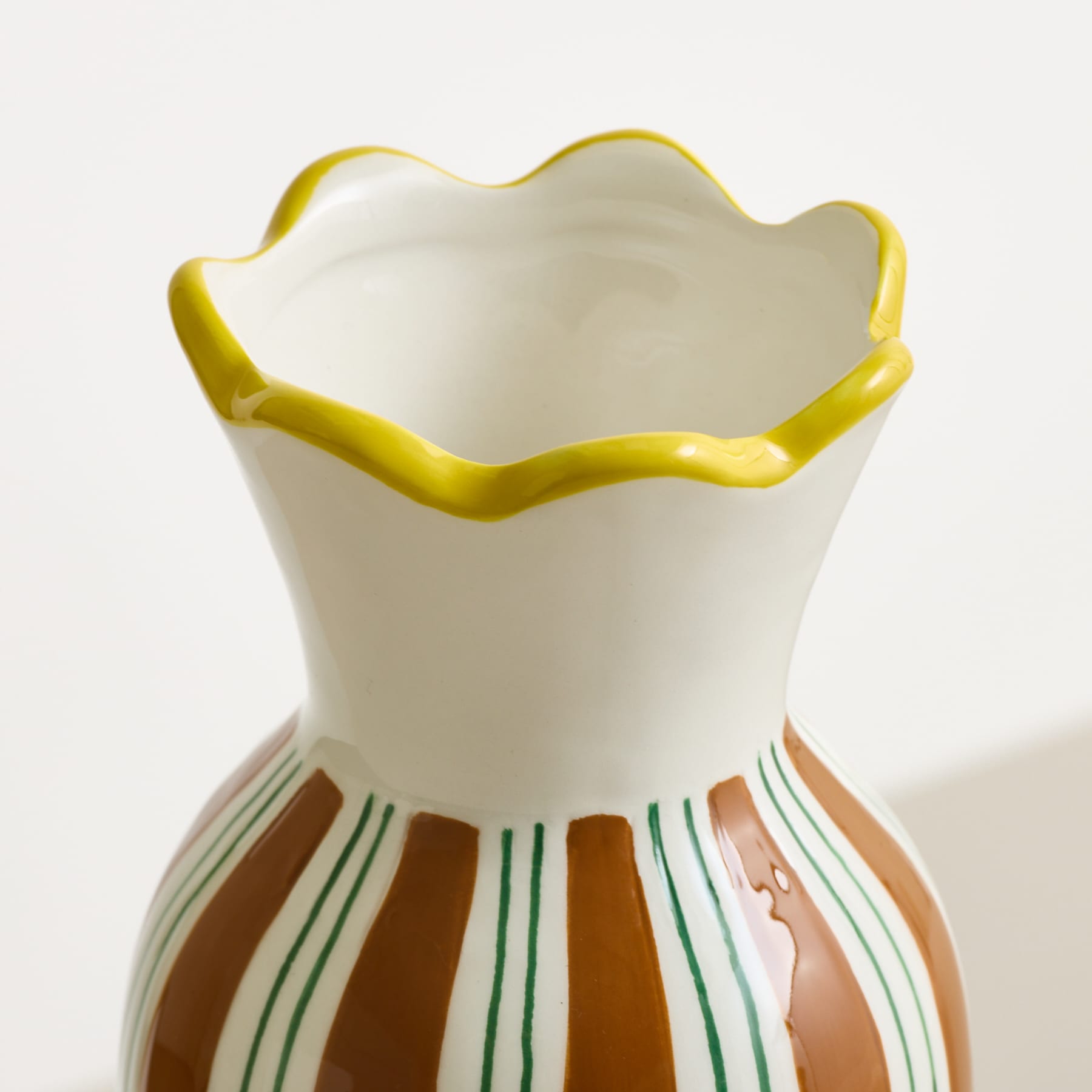 Eine weiße Keramikvase mit gelbem, gerüschtem Rand und braunen und grünen Streifen steht vor einem schlichten weißen Hintergrund.