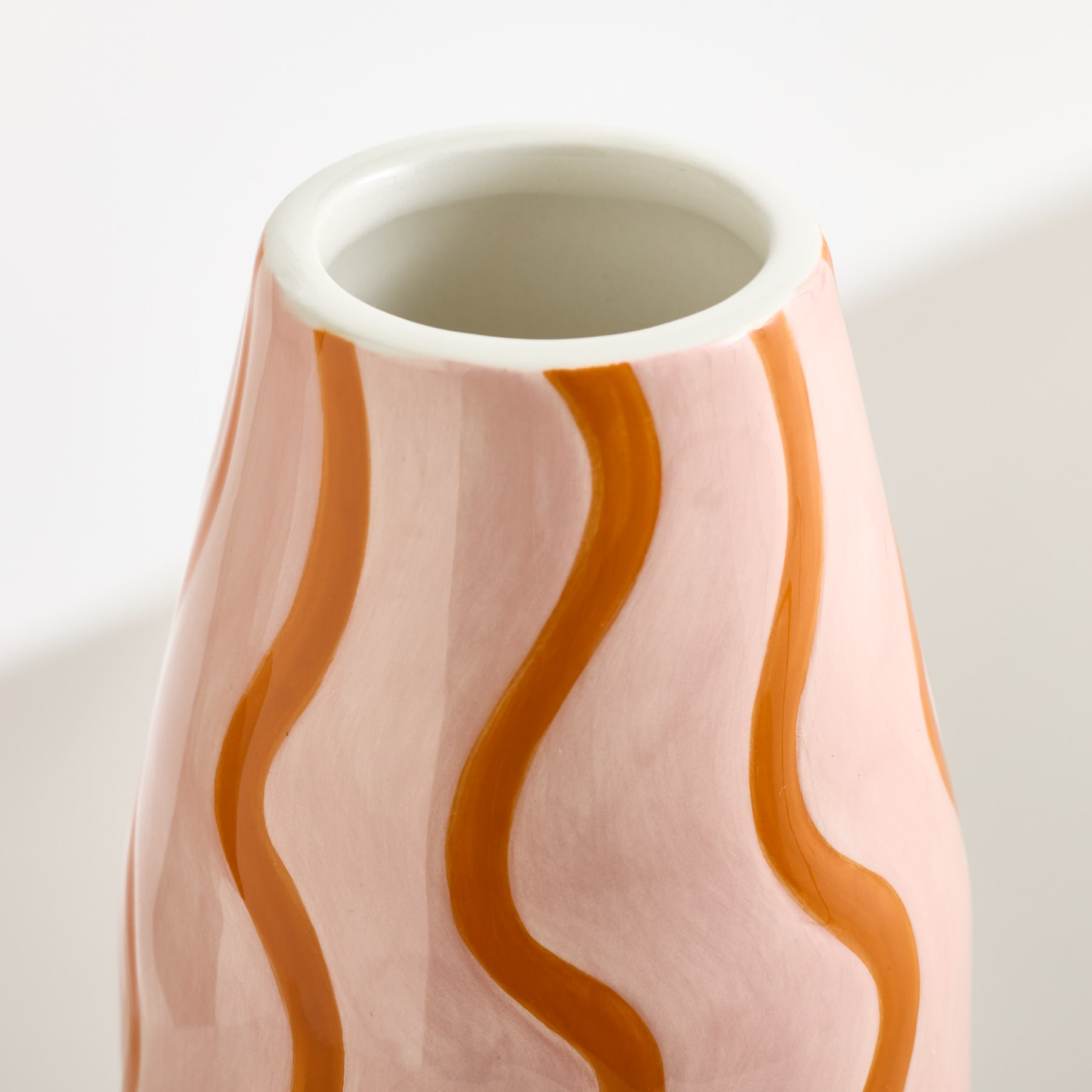 Im Vordergrund steht eine rosa Vase mit gewellten braunen Streifen, vor einem sanften weißen Hintergrund, der einen Schatten wirft.