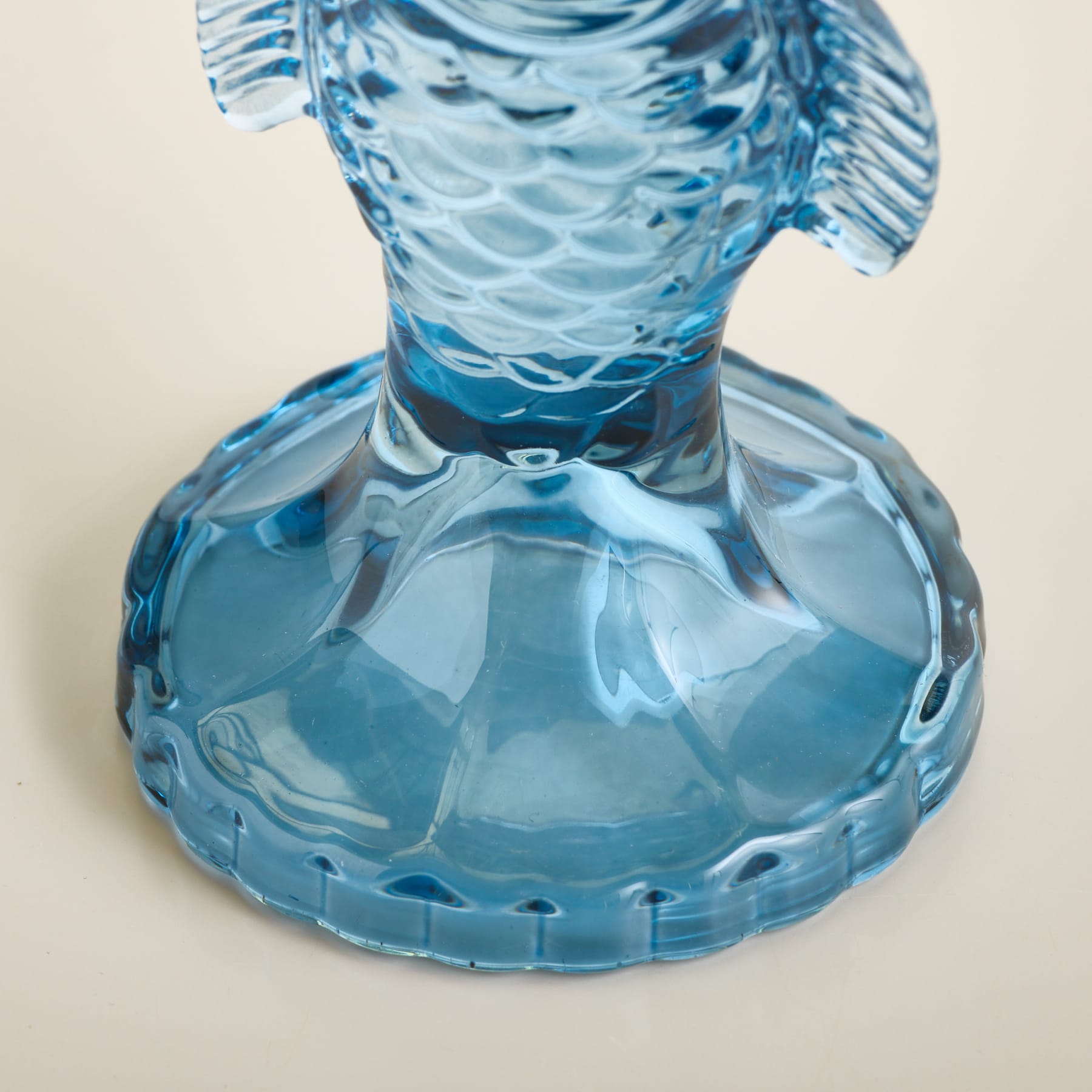 Eine blaue Glasfischfigur mit strukturierter Unterseite steht auf einer schlichten, hellen Oberfläche.