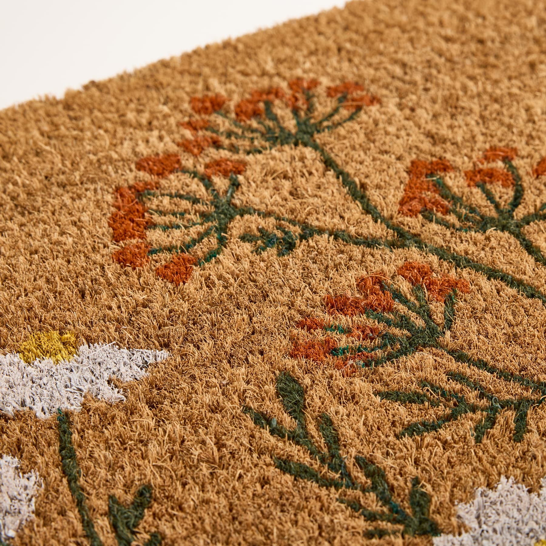 Nahaufnahme einer Kokosfaser-Fußmatte mit einem floralen Muster aus orangefarbenen Wildblumen und weißen Gänseblümchen im Vordergrund vor einem schlichten weißen Hintergrund.