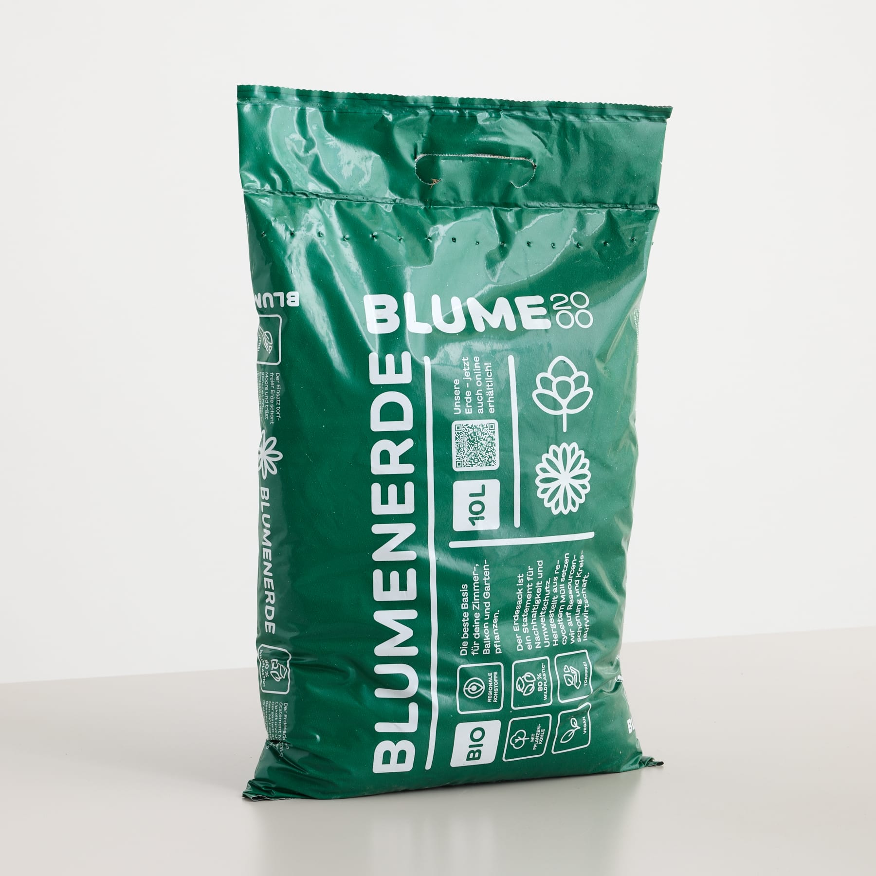 Ein grüner Sack mit „BLUMENERDE“-Erde steht auf einer hellgrauen Fläche vor einem schlichten weißen Hintergrund.