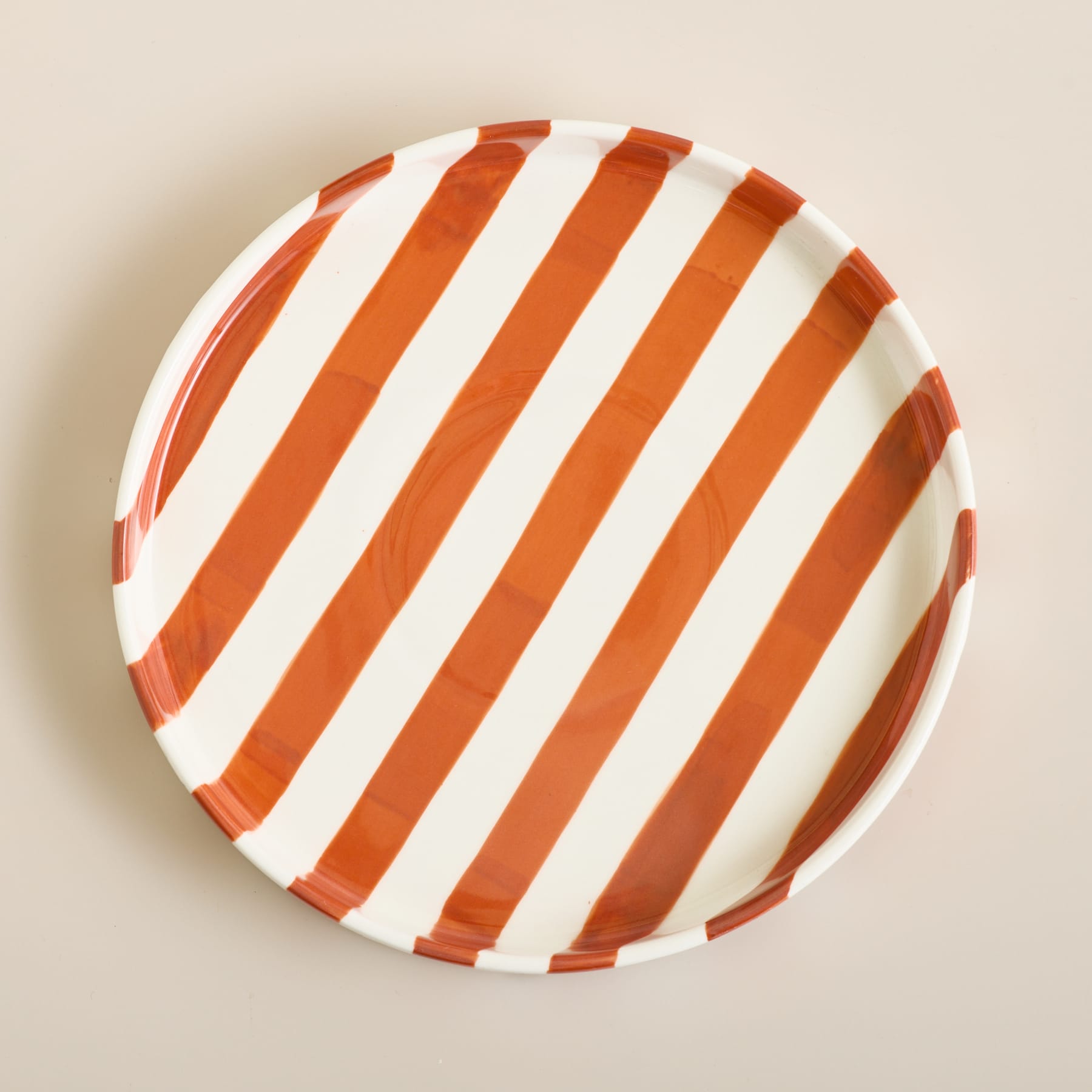 Ein runder Keramikteller mit diagonalen orangefarbenen und weißen Streifen steht auf einer schlichten, hellbeigen Oberfläche.
