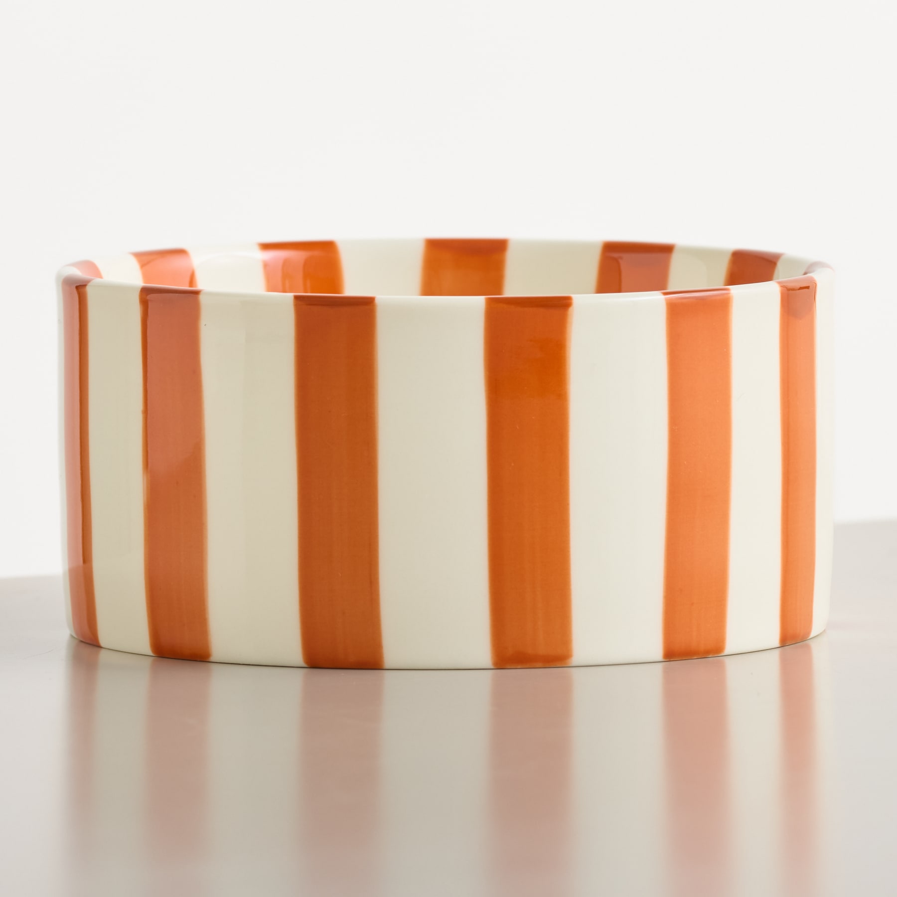 Eine cremefarbene und orange gestreifte Keramikschale steht auf einer reflektierenden grauen Oberfläche vor einem schlichten weißen Hintergrund.